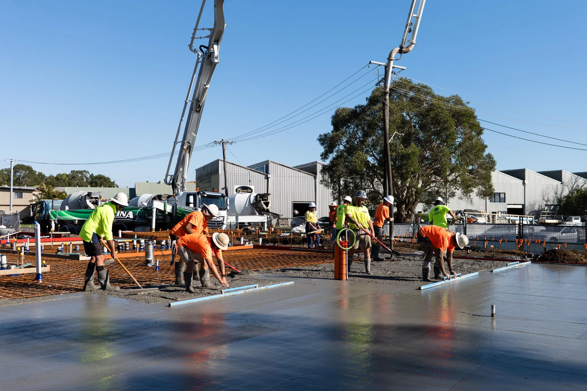 Construction workers in bright clothing and hard hats are pouring and leveling concrete on a building site. Heavy machinery, power lines, and industrial buildings are visible in the background, with a large tree to the right.