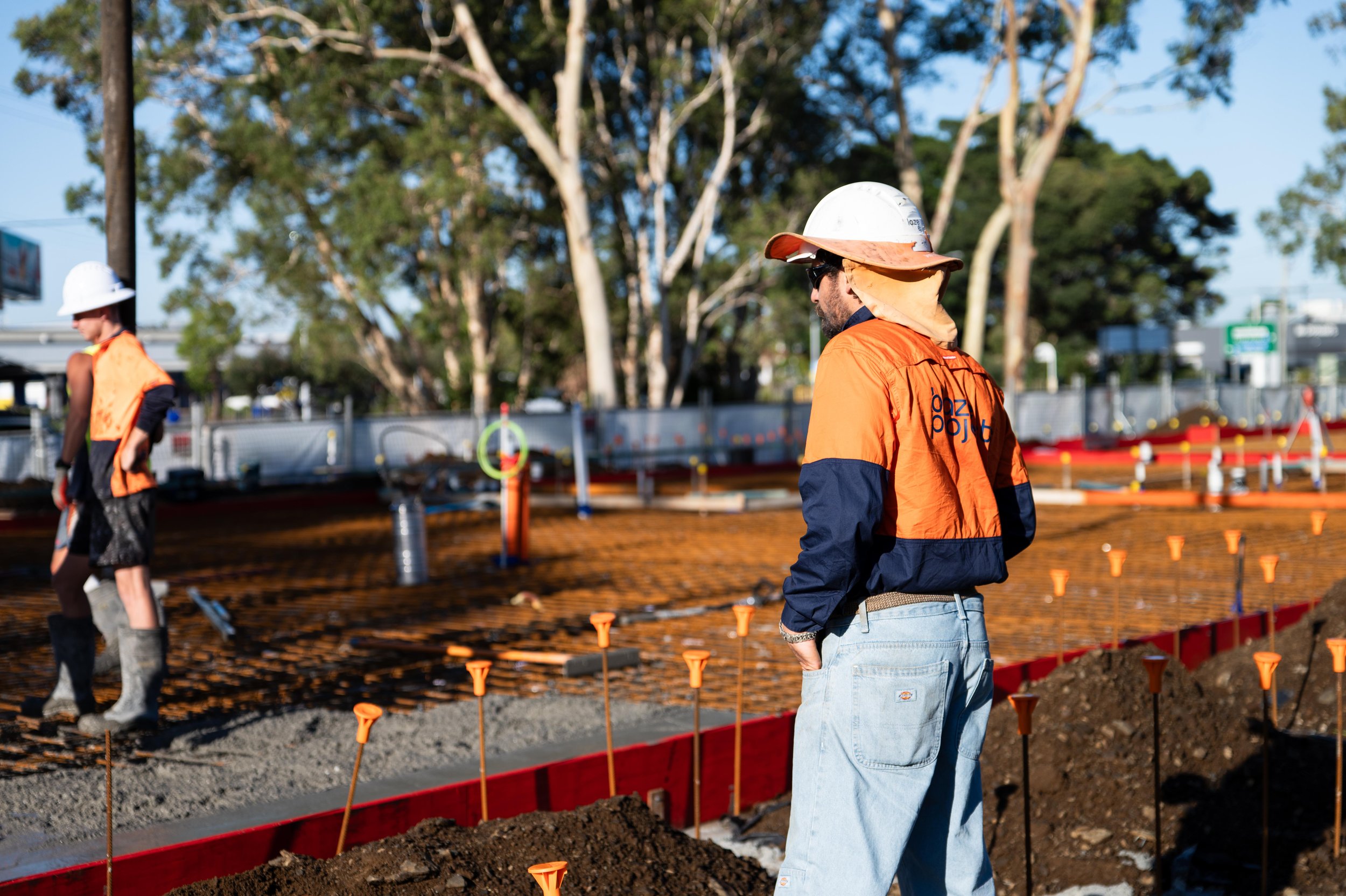 Construction workers in safety gear working on a site with trees in the background. One worker is watching while another is working on the ground.