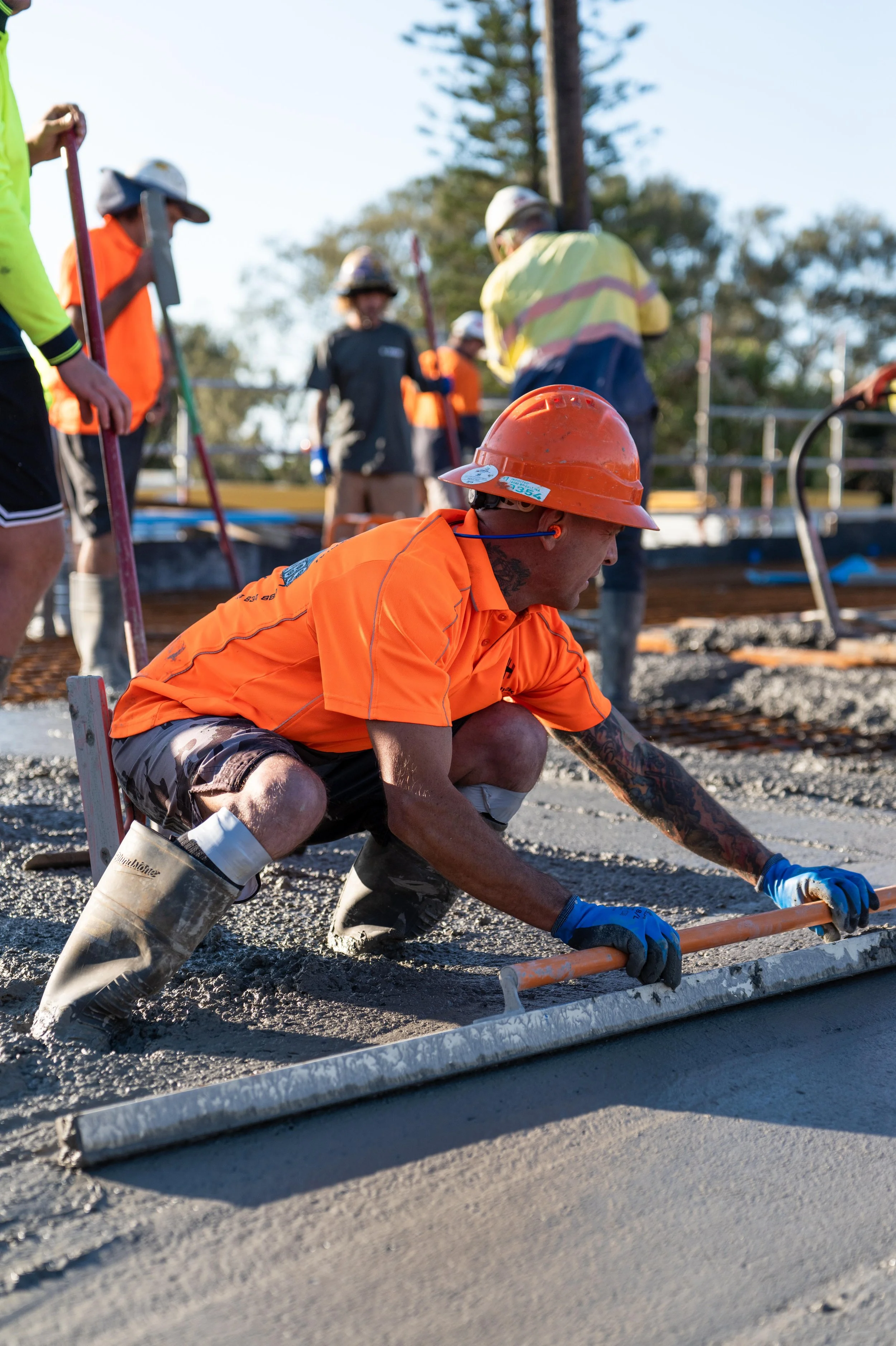 A construction worker in an orange shirt and hard hat kneels on the ground smoothing wet concrete with a long tool, while other workers in safety gear work in the background.