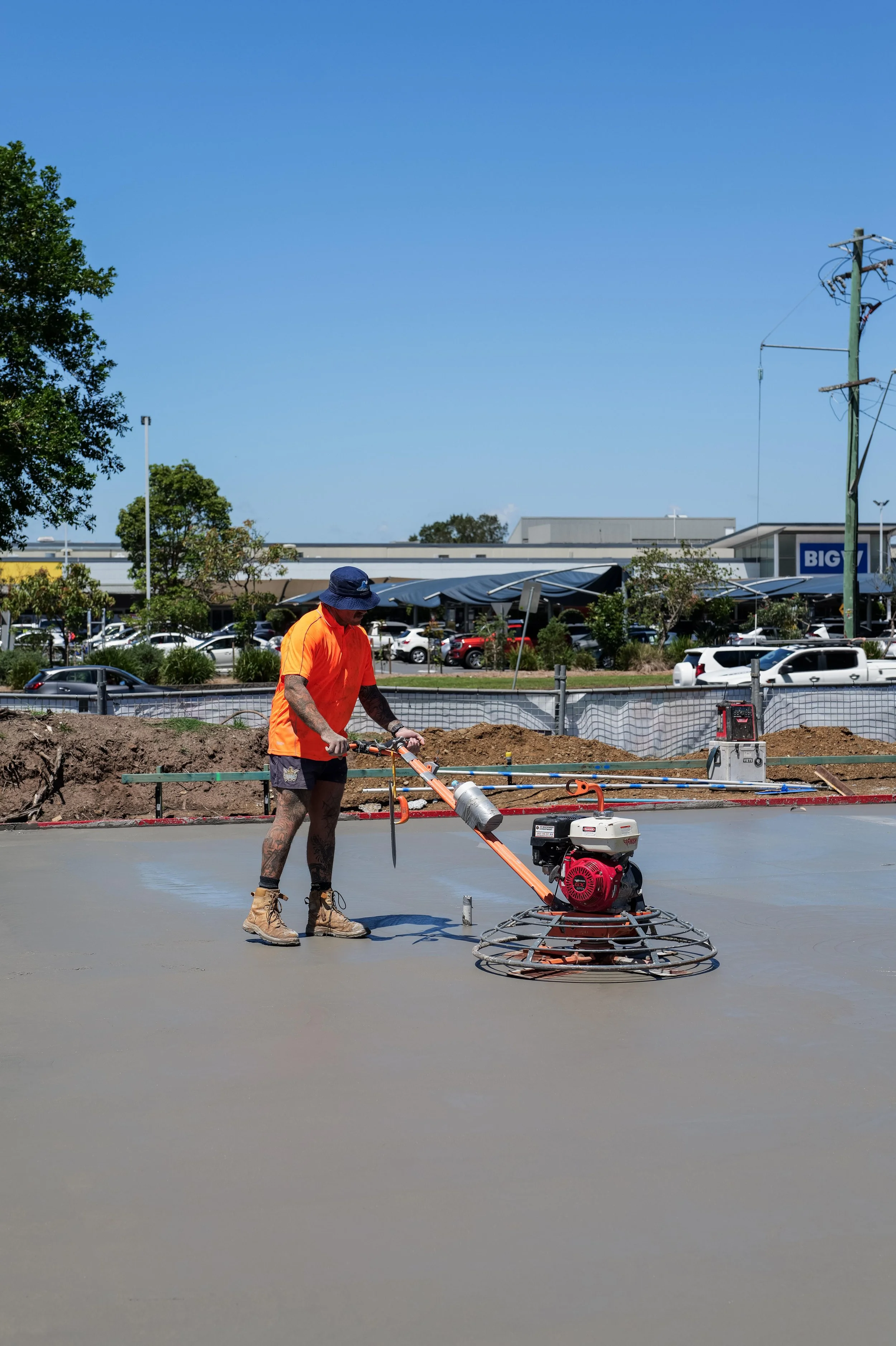 Worker in orange shirt and blue hat smoothing freshly poured concrete with a power trowel at a construction site, with a shopping center and parked cars in the background.
