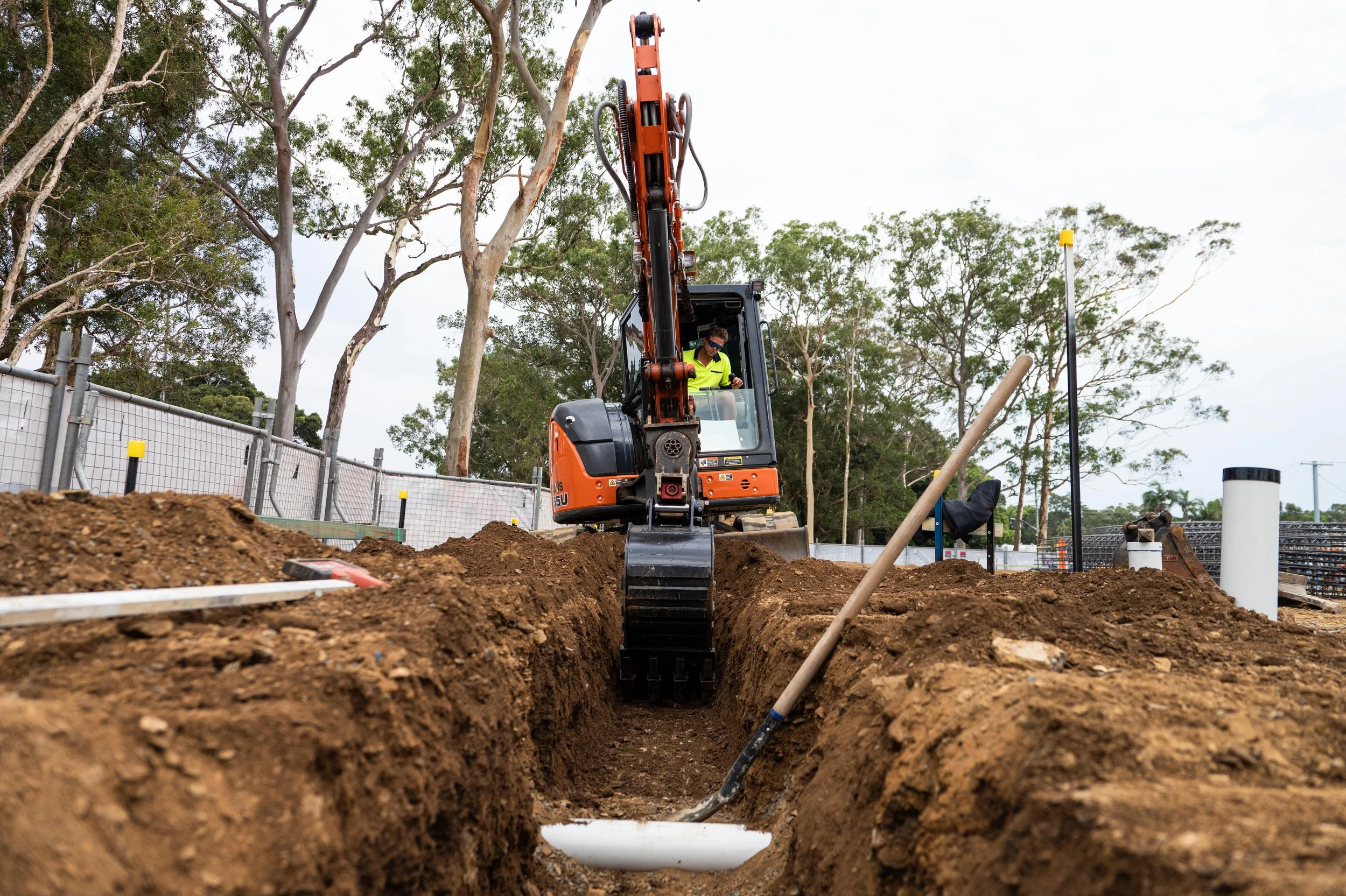 A construction worker operating an orange excavator in a trench, digging the ground during a construction project outdoors with trees and a fence in the background.