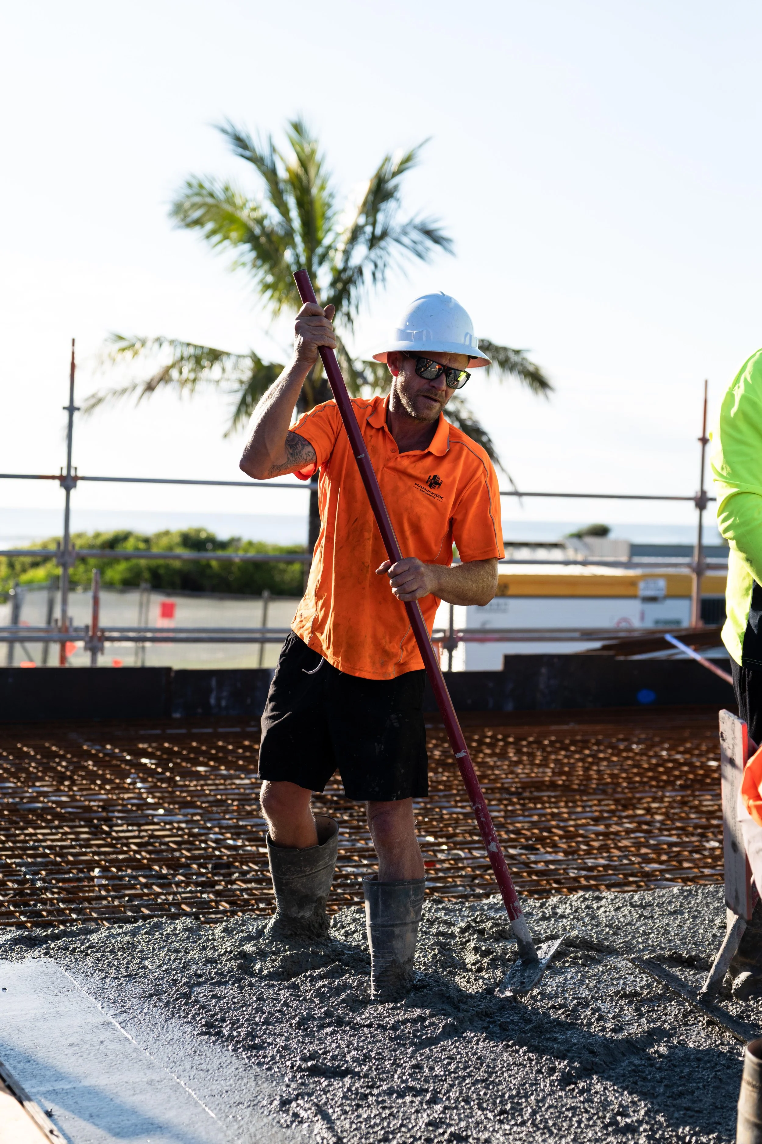 A construction worker in an orange shirt, black shorts, and tall boots is pouring concrete with a long-handled tool on a construction site outdoors, with palm trees and a bright sky in the background.