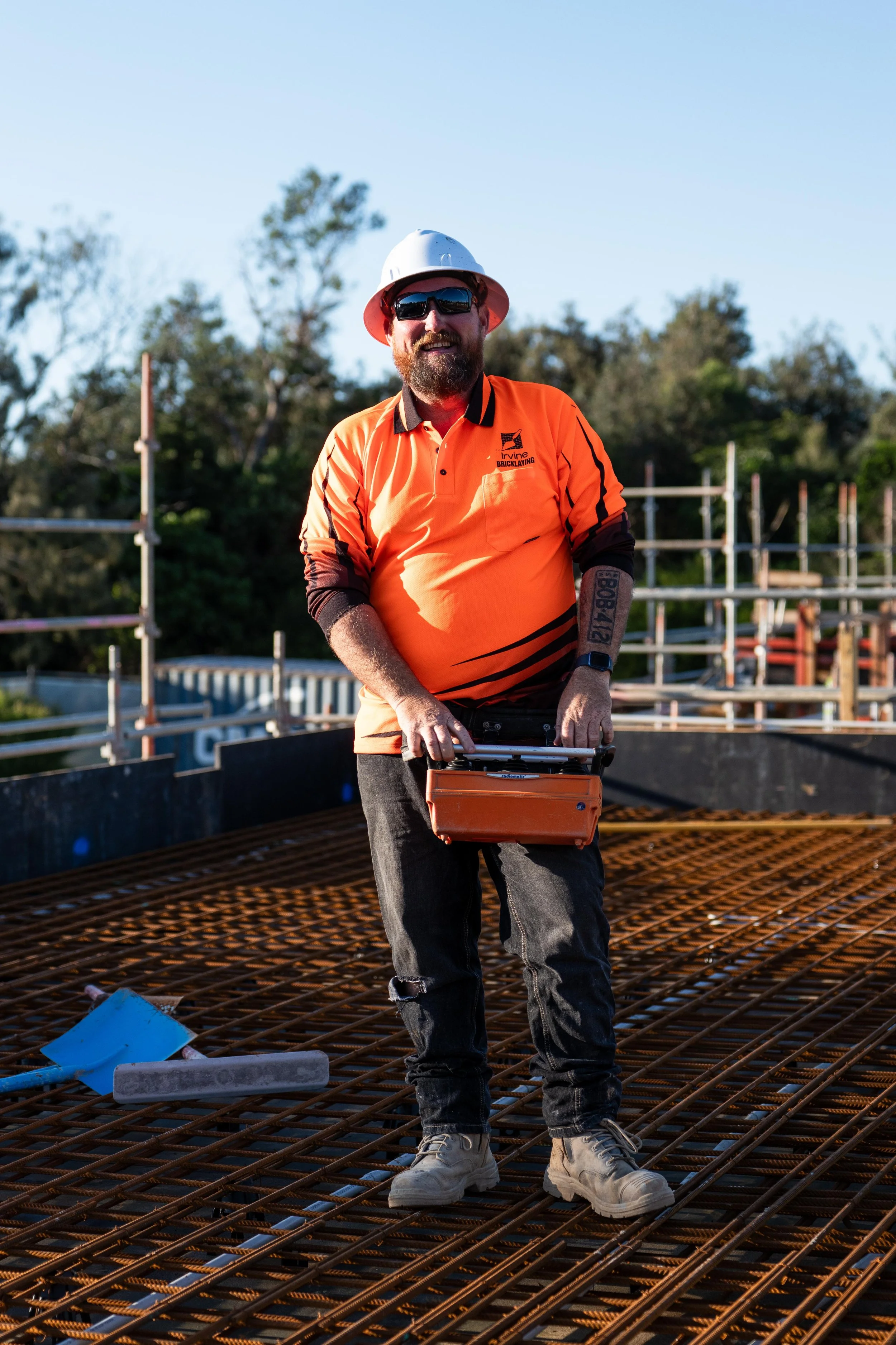 A construction worker standing on a steel rebar grid at a construction site, wearing an orange shirt, hard hat, sunglasses, and holding a toolbox.