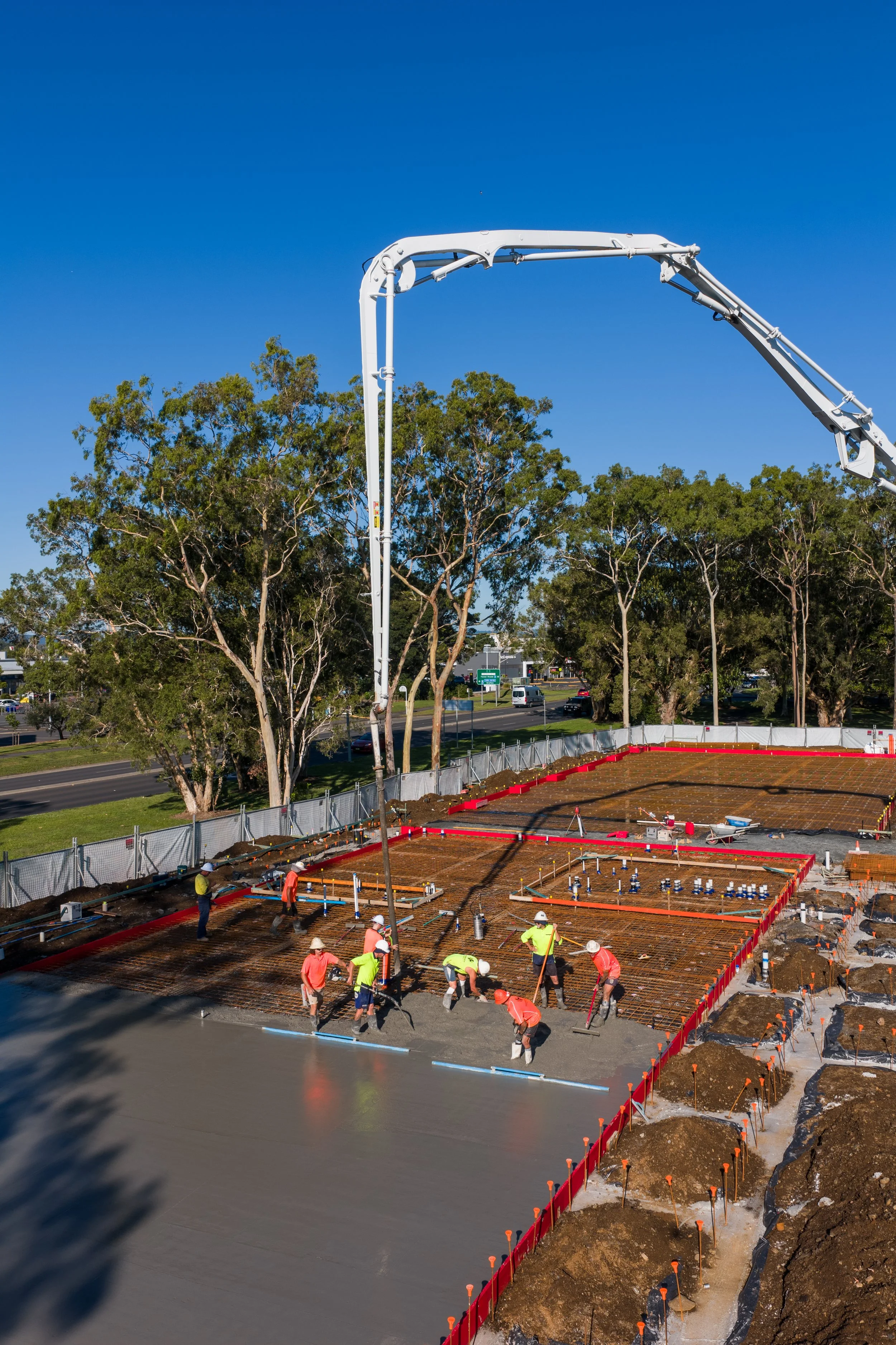 Construction workers pouring concrete on a building foundation with a concrete pump truck overhead, trees and a road in the background.