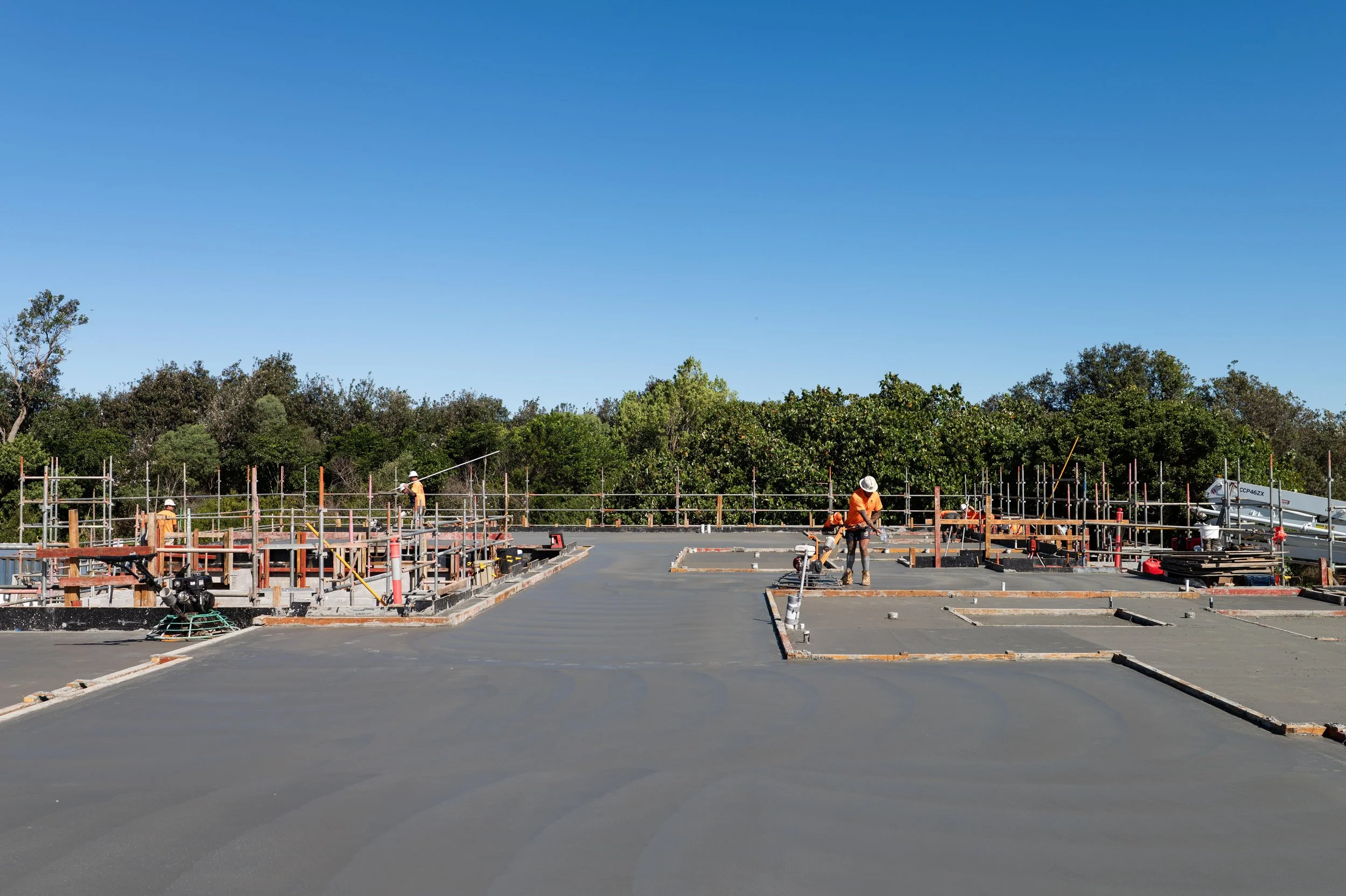 Construction workers at a building site spreading and smoothing concrete on a flat surface with trees in the background.
