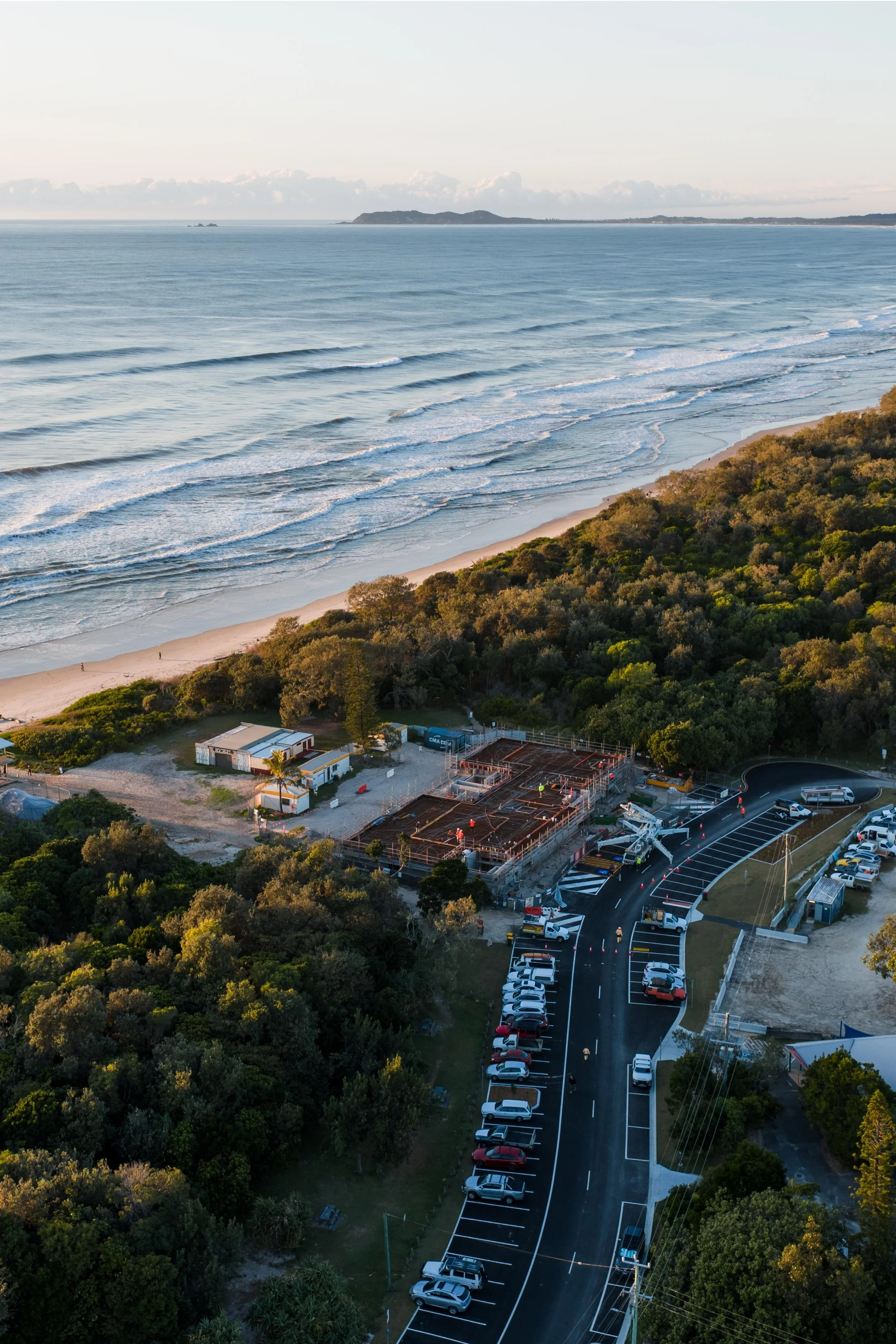 Aerial view of a construction site near a beach, with parked cars and greenery