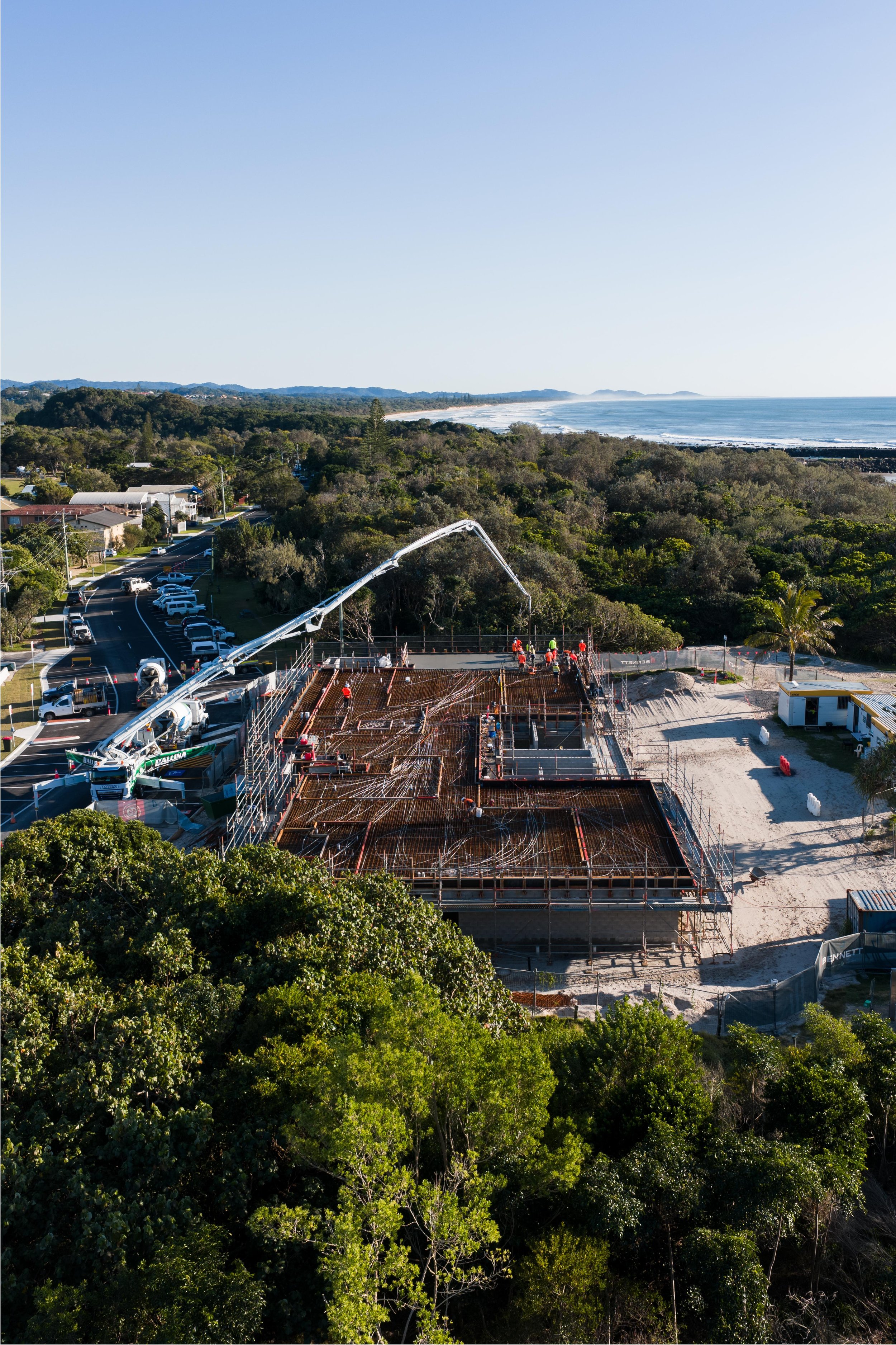 Construction site with concrete pouring activity near a forest and beach, under a clear sky.