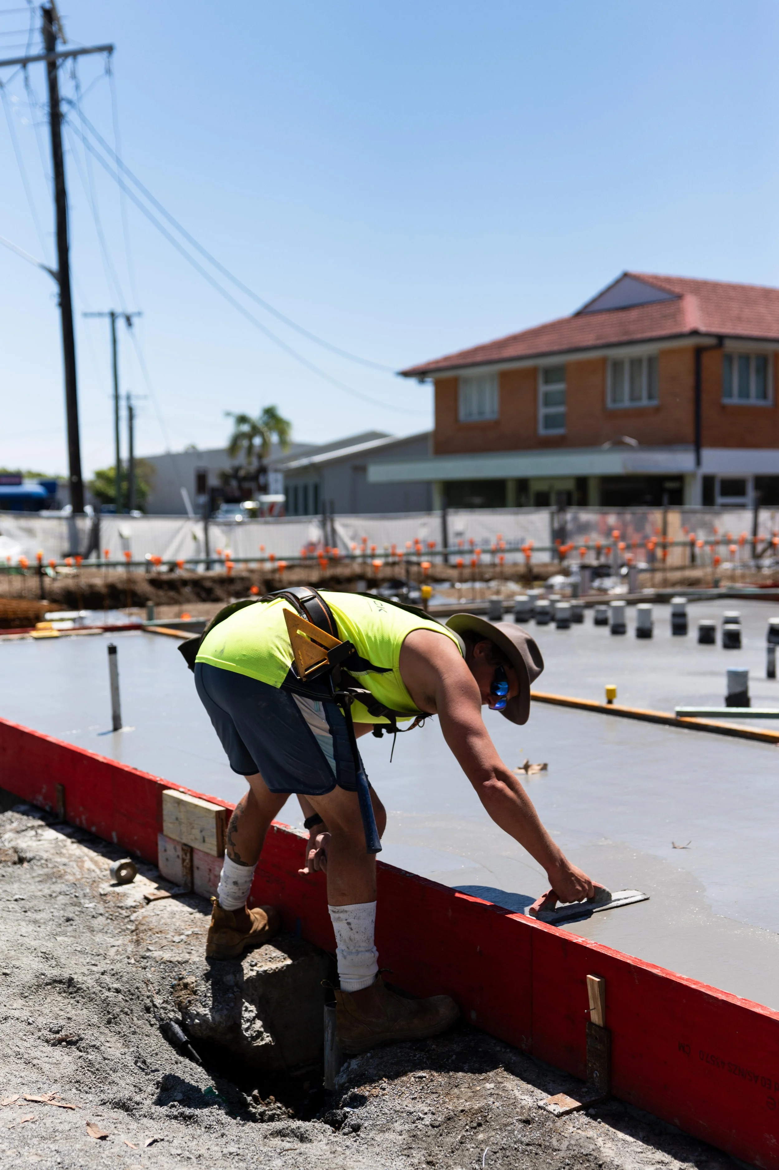 A construction worker in a yellow vest, hat, and sunglasses smoothing wet concrete on a sidewalk at a construction site in a residential area.