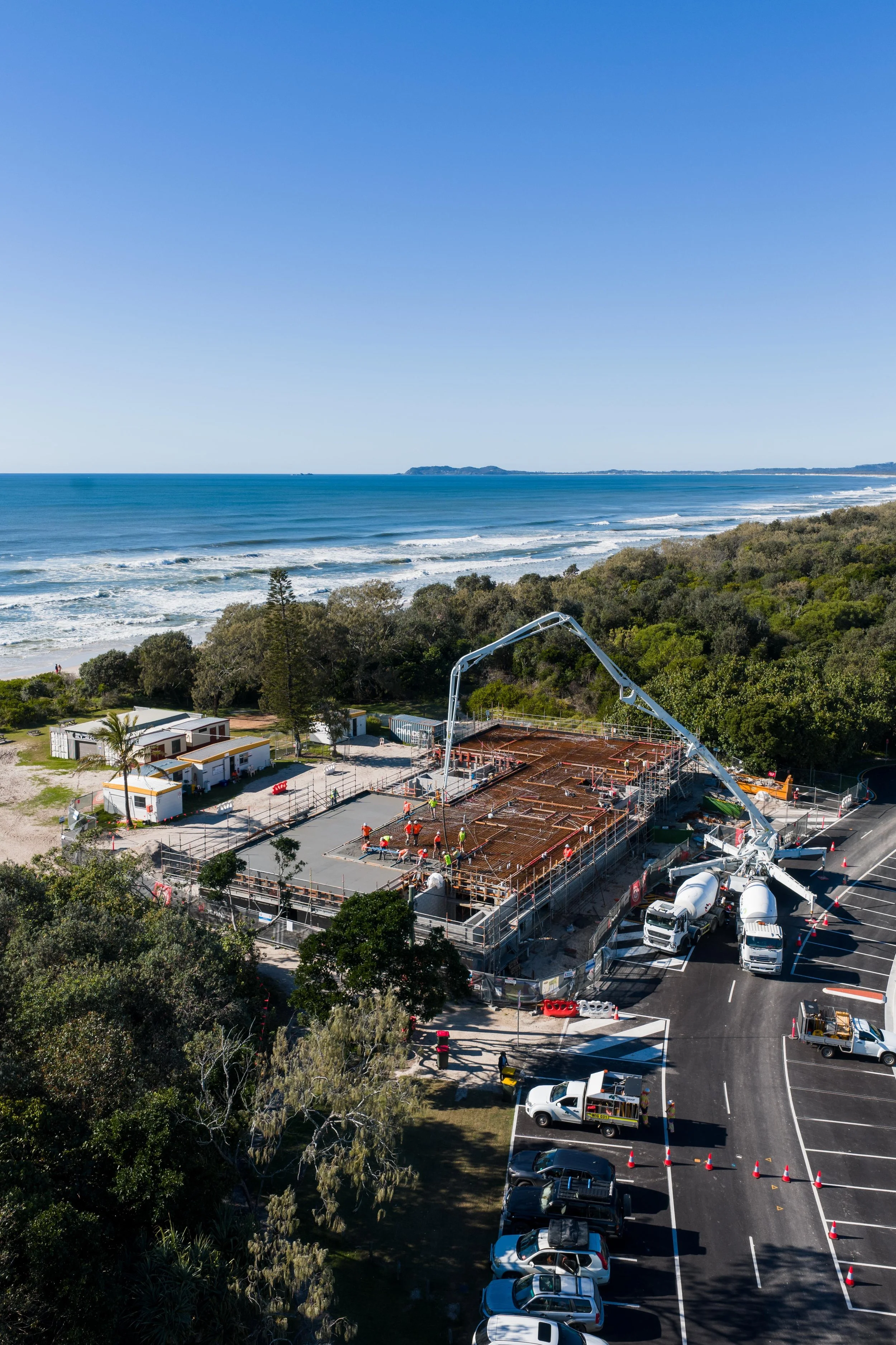 Construction site with workers pouring concrete for a building near a beach, with a parking lot and lush greenery in the background.