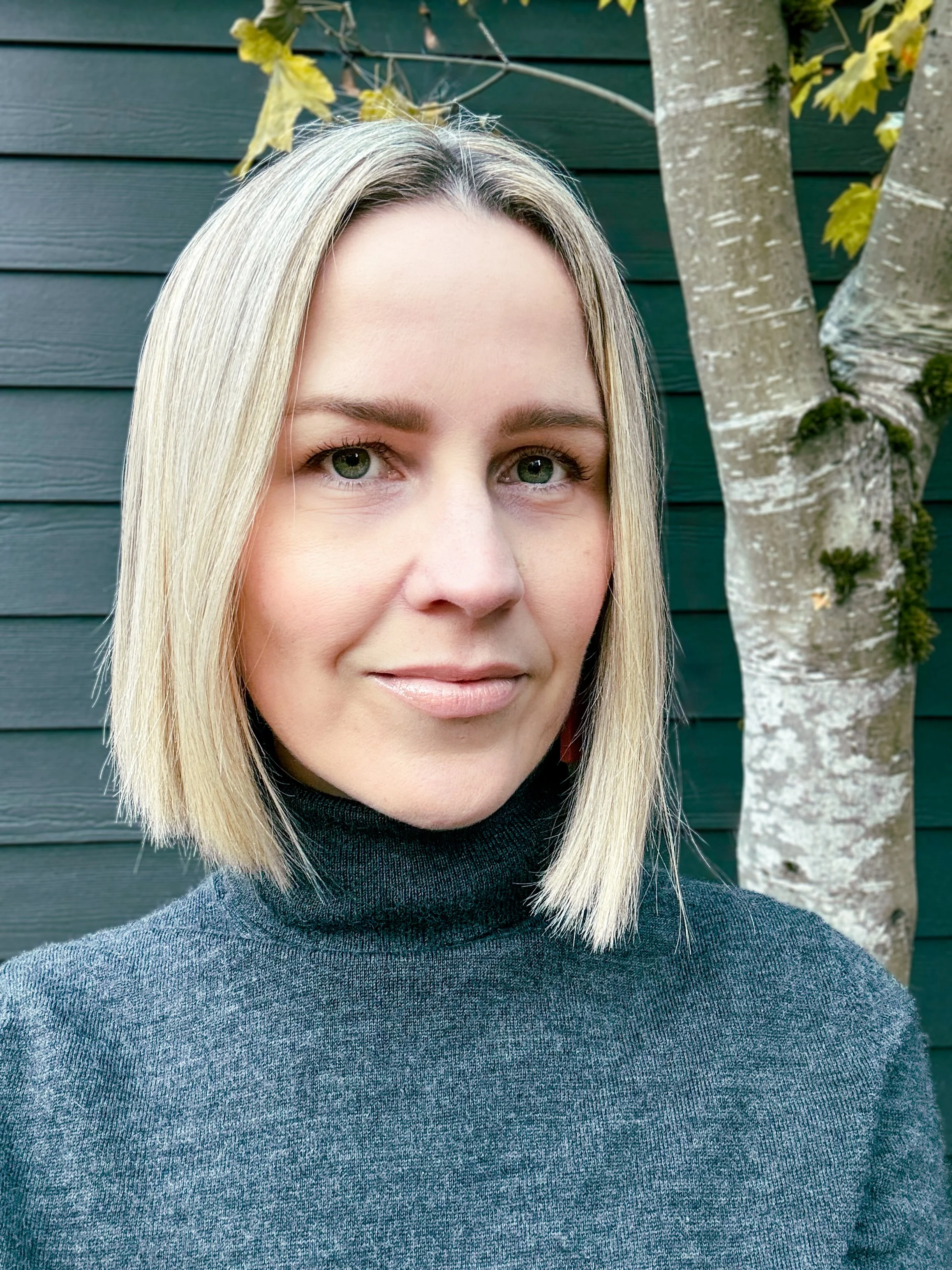 A woman with blonde hair and green eyes wearing a dark gray turtleneck sweater standing outdoors in front of a dark green wooden wall and a tree with white bark and green leaves.