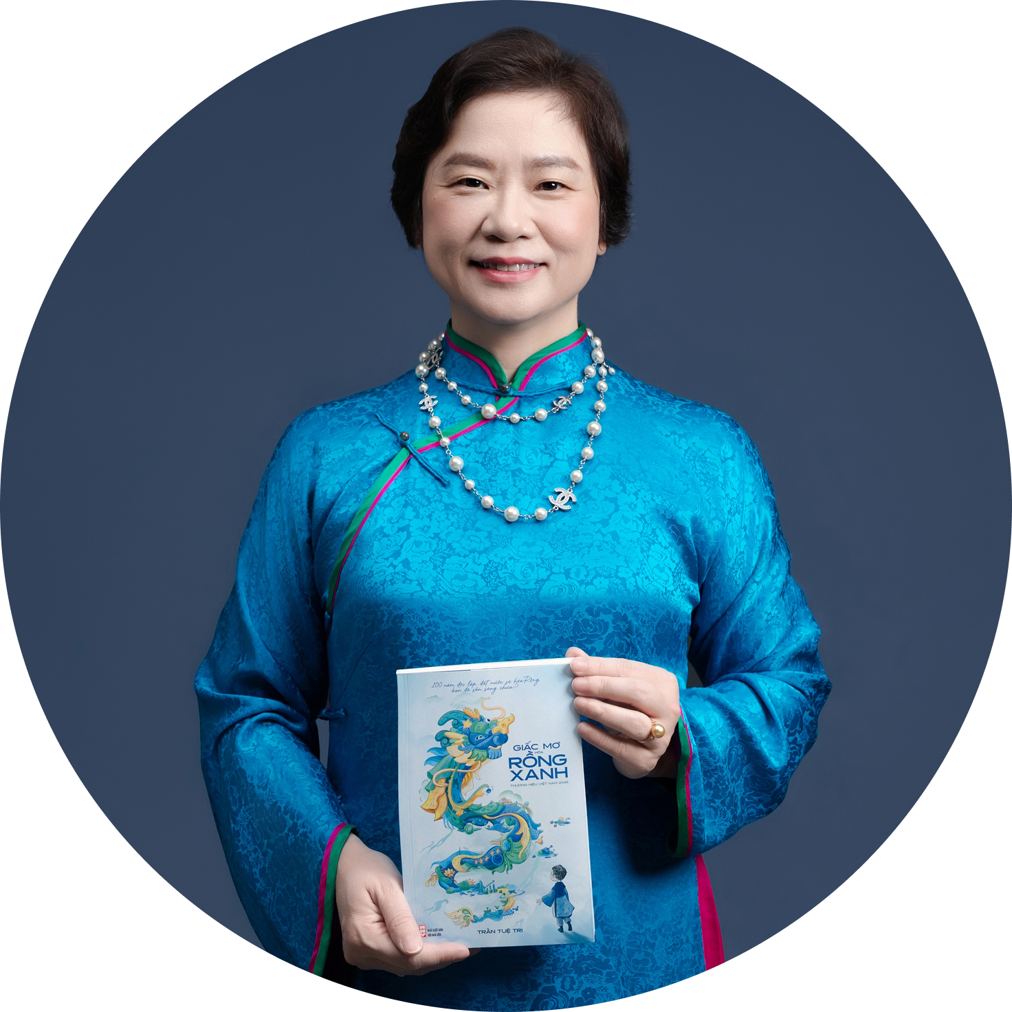 A woman wearing a traditional blue Vietnamese áo dài, holding a book with a dragon illustration on the cover, standing in front of a gray background.