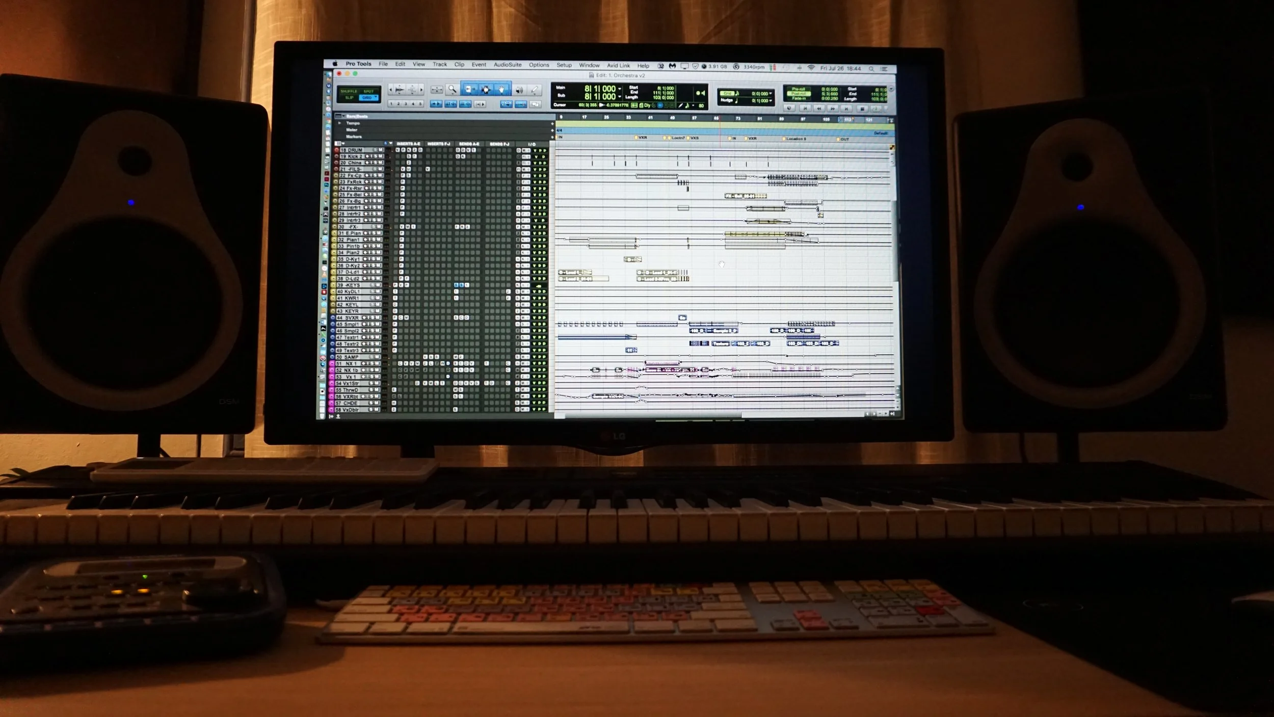 Computer monitor displaying music production software, surrounded by studio speakers, a keyboard, and controller on a desk.