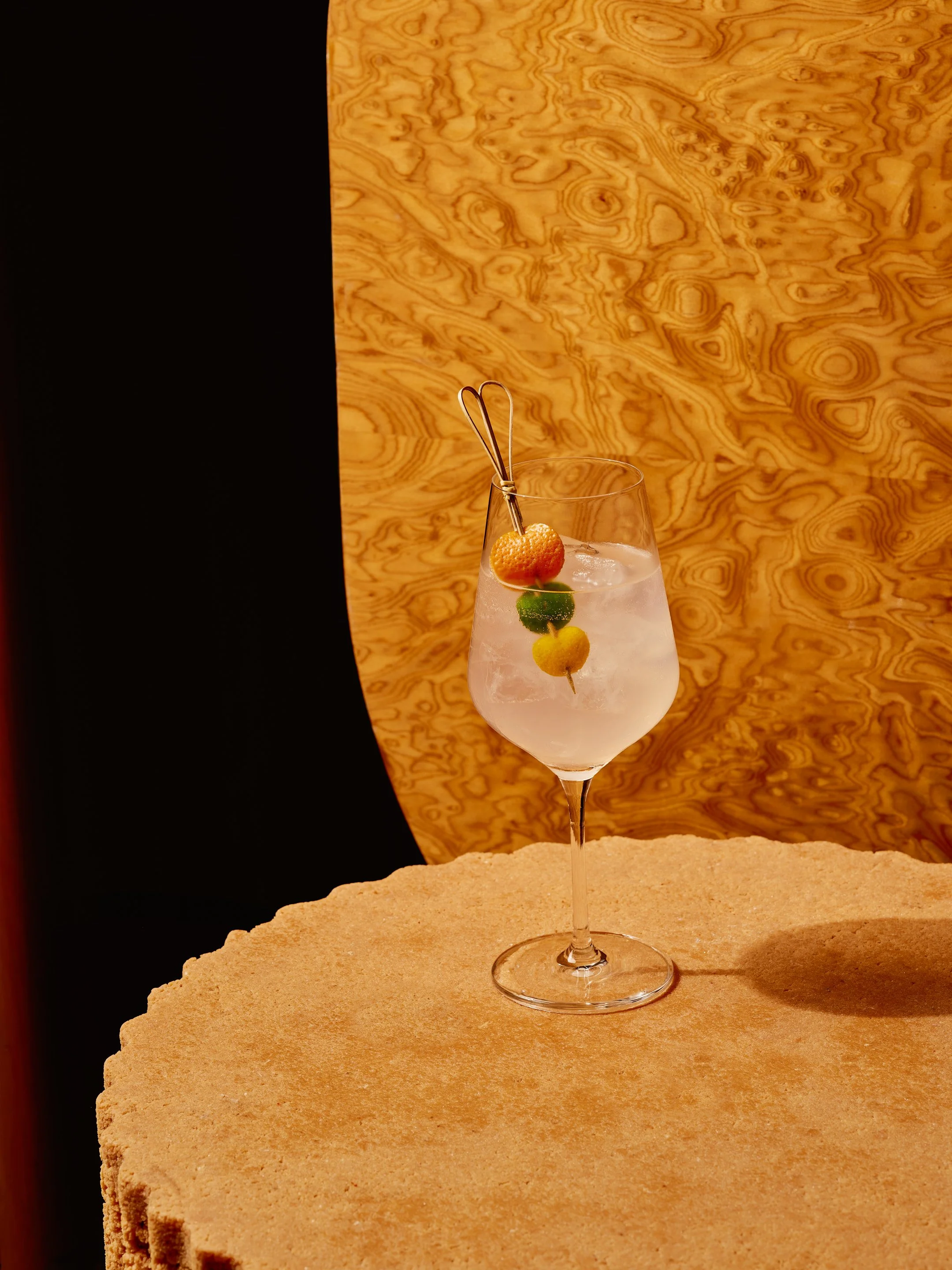A cocktail glass with a skewered garnish of a cherry, lime, and lemon slices, filled with a clear beverage and ice cubes, set on a textured beige surface with a wood-paneled background.