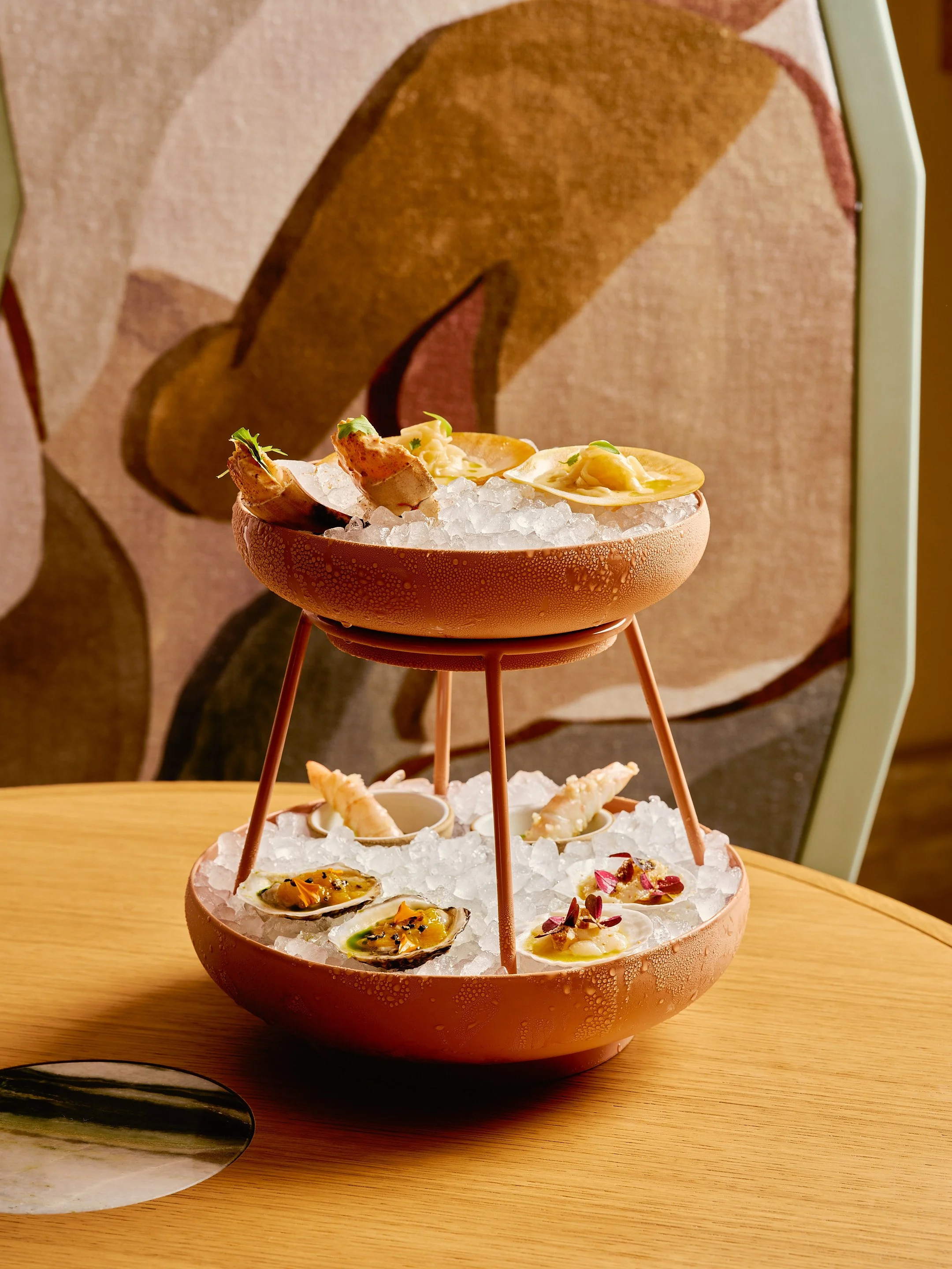 An elegant food display featuring a two-tiered copper and stone serving stand filled with oysters on a bed of ice.