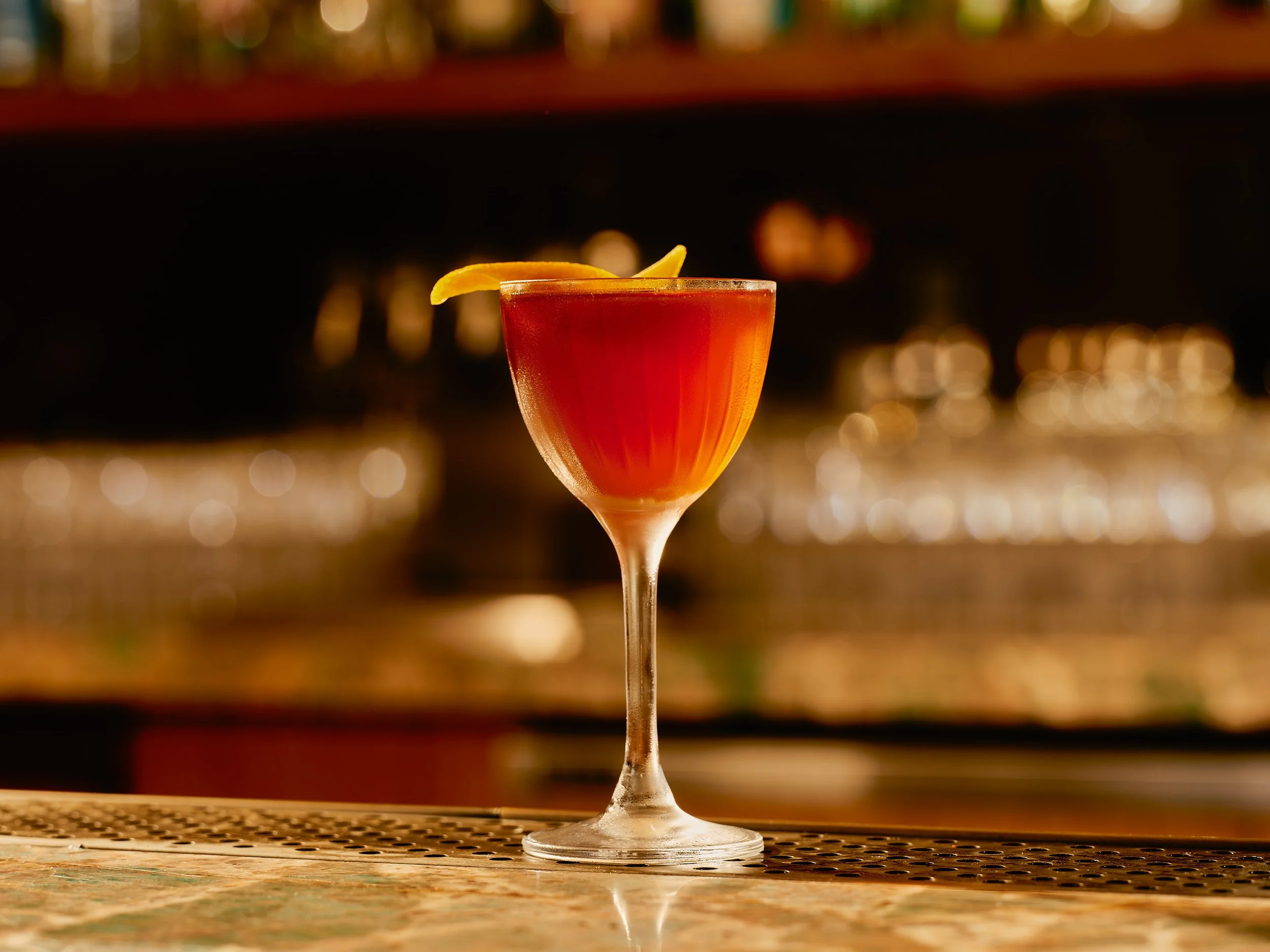 A cocktail glass with a red drink and a lemon wedge garnish, placed on a bar counter with blurred bar background.