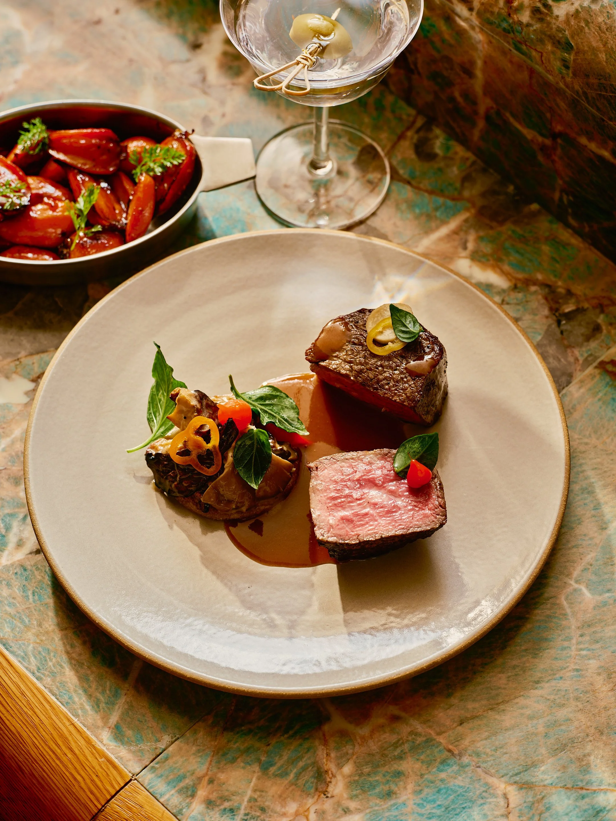 Plate with three pieces of cooked beef, garnished with herbs and vegetables, placed on a textured surface. In the background, a glass of water with a lemon wedge, and a bowl of glazed cherry tomatoes garnished with herbs.