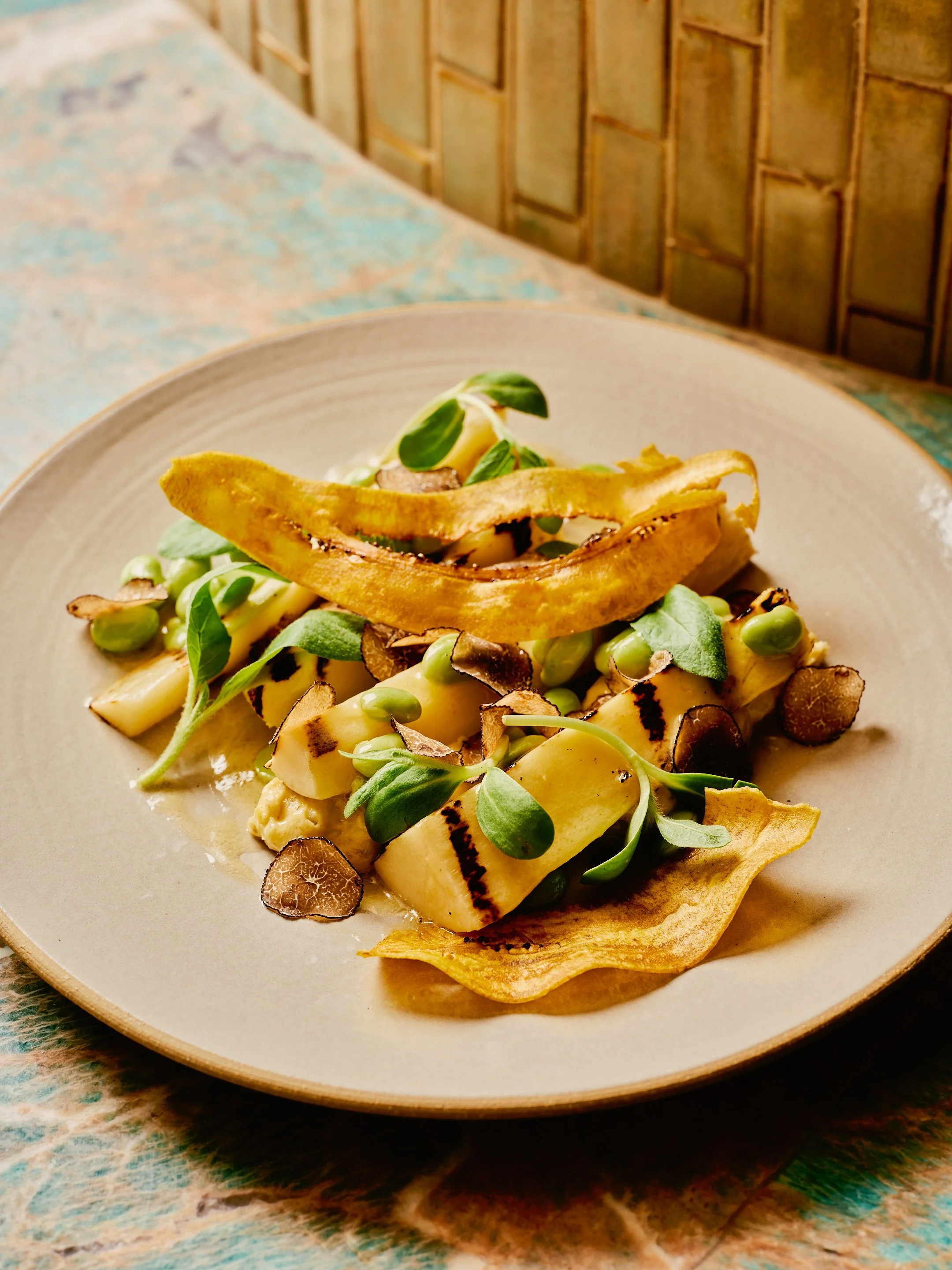 A plated dish with grilled vegetables, microgreens, and crispy bread strips on a beige plate.