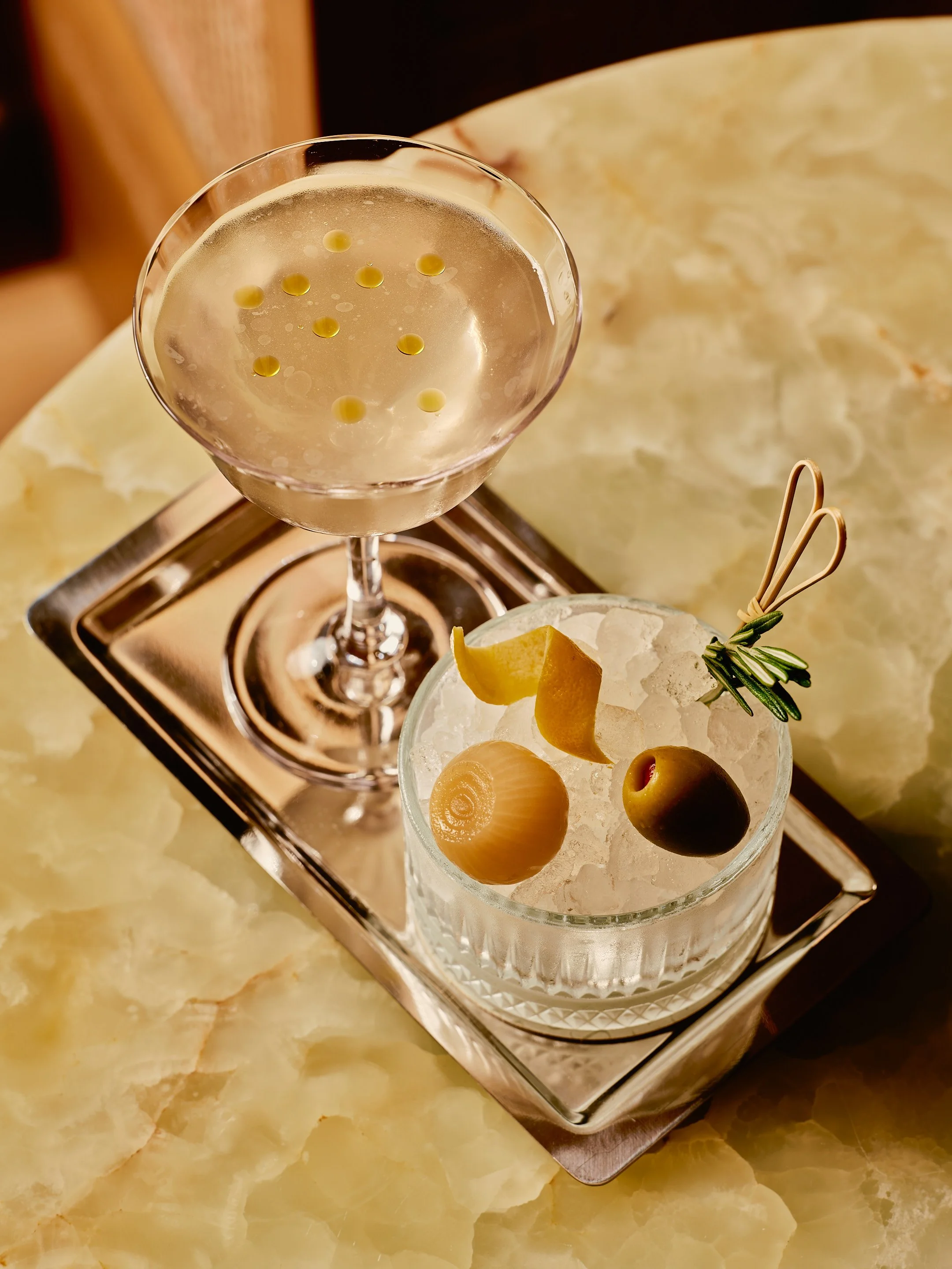 A cocktail glass with a clear drink and yellow dots inside, placed on a metal tray. Next to it is a short glass filled with ice and garnished with olives, lemon peel, and a sprig of rosemary, also on a metal tray.
