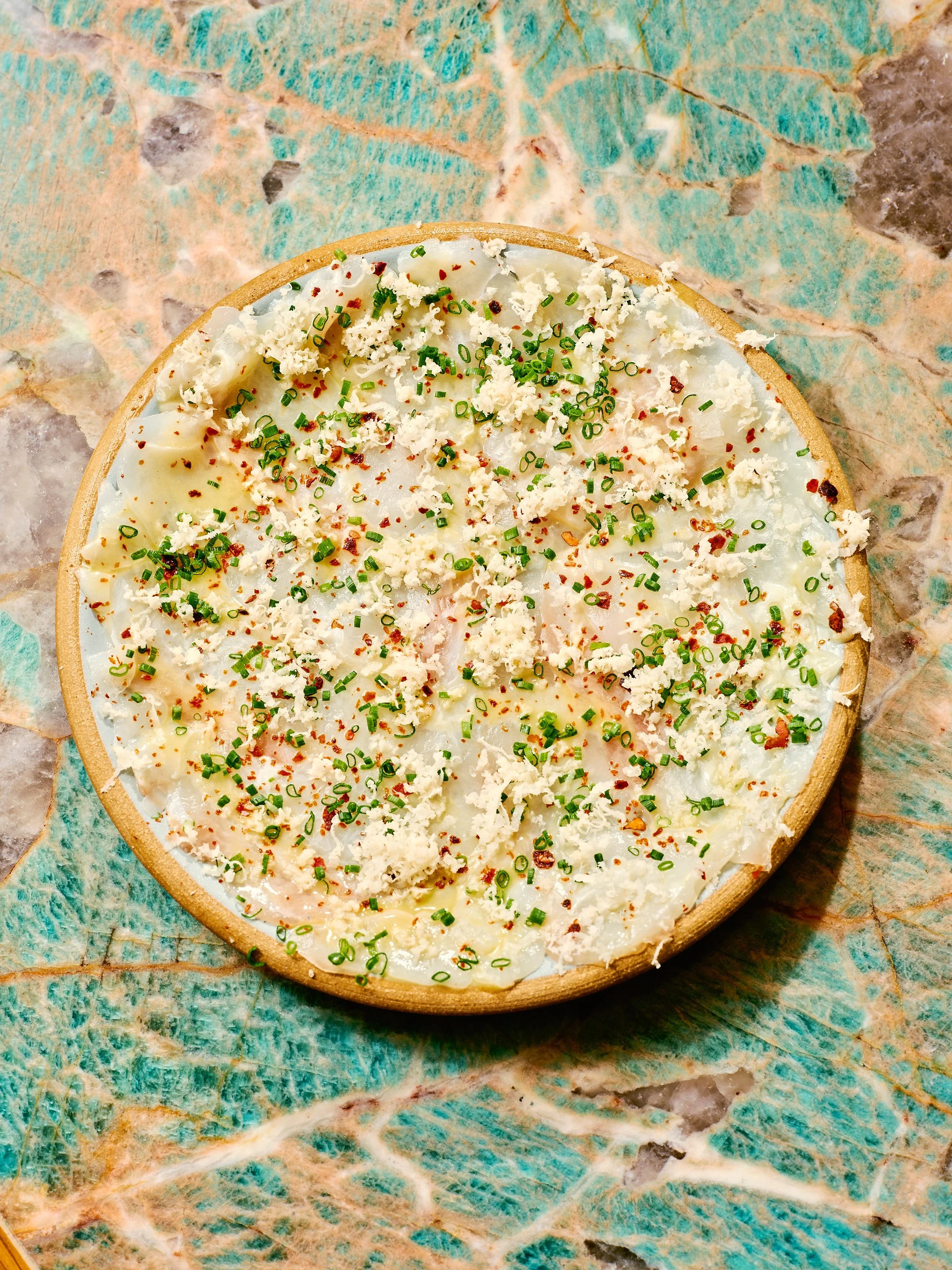 A plate of sliced cheese topped with chopped chives, red pepper flakes, and grated cheese on a decorative surface.