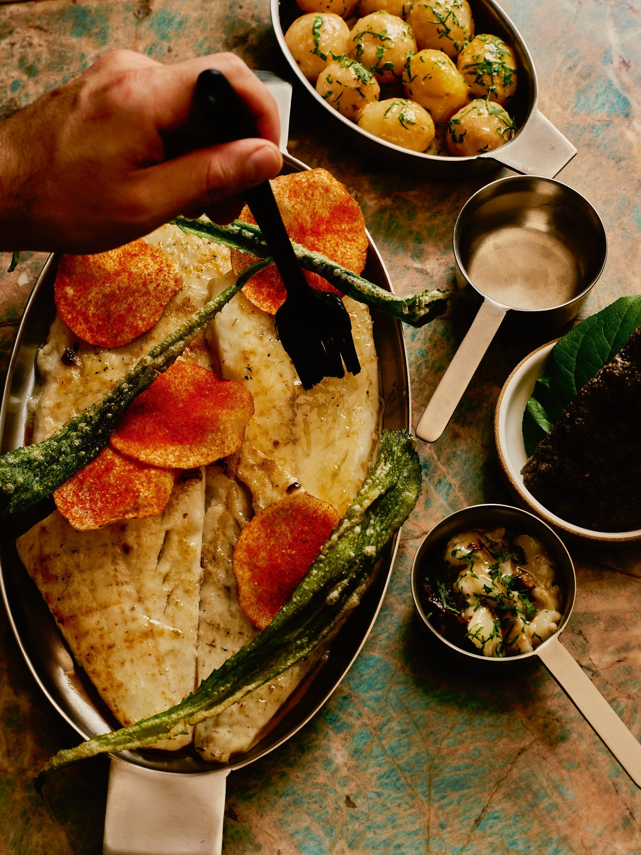 A person is using a fork to serve a piece of fish topped with orange-colored crispy garnishes, with green vegetables on the fish. Surrounding the main dish are bowls of boiled potatoes, a bowl of seaweed-wrapped rice, and a small bowl of potato salad