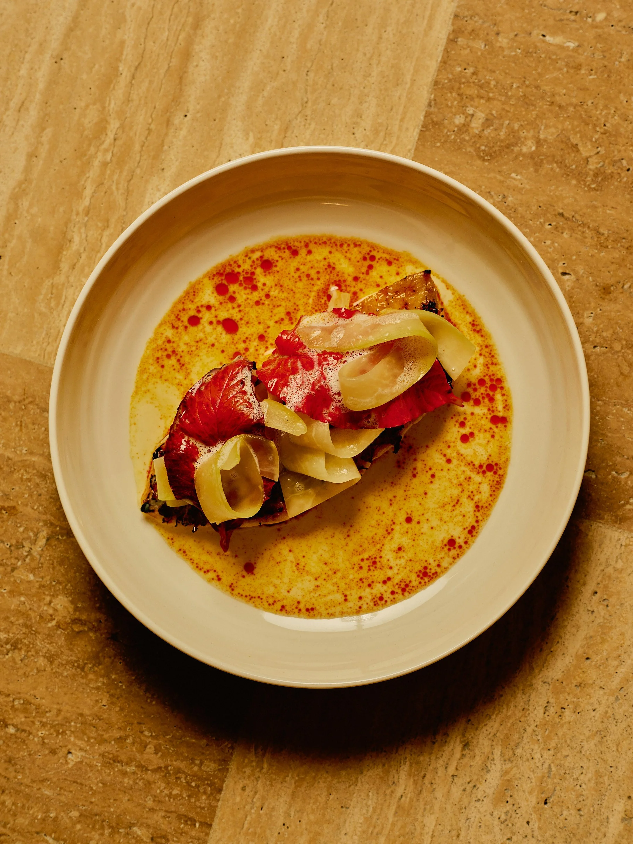 A white bowl containing a colorful, gourmet dish with a roasted vegetable topped with pickled vegetables and surrounded by a yellowish sauce with red oil droplets, placed on a multi-colored stone countertop.