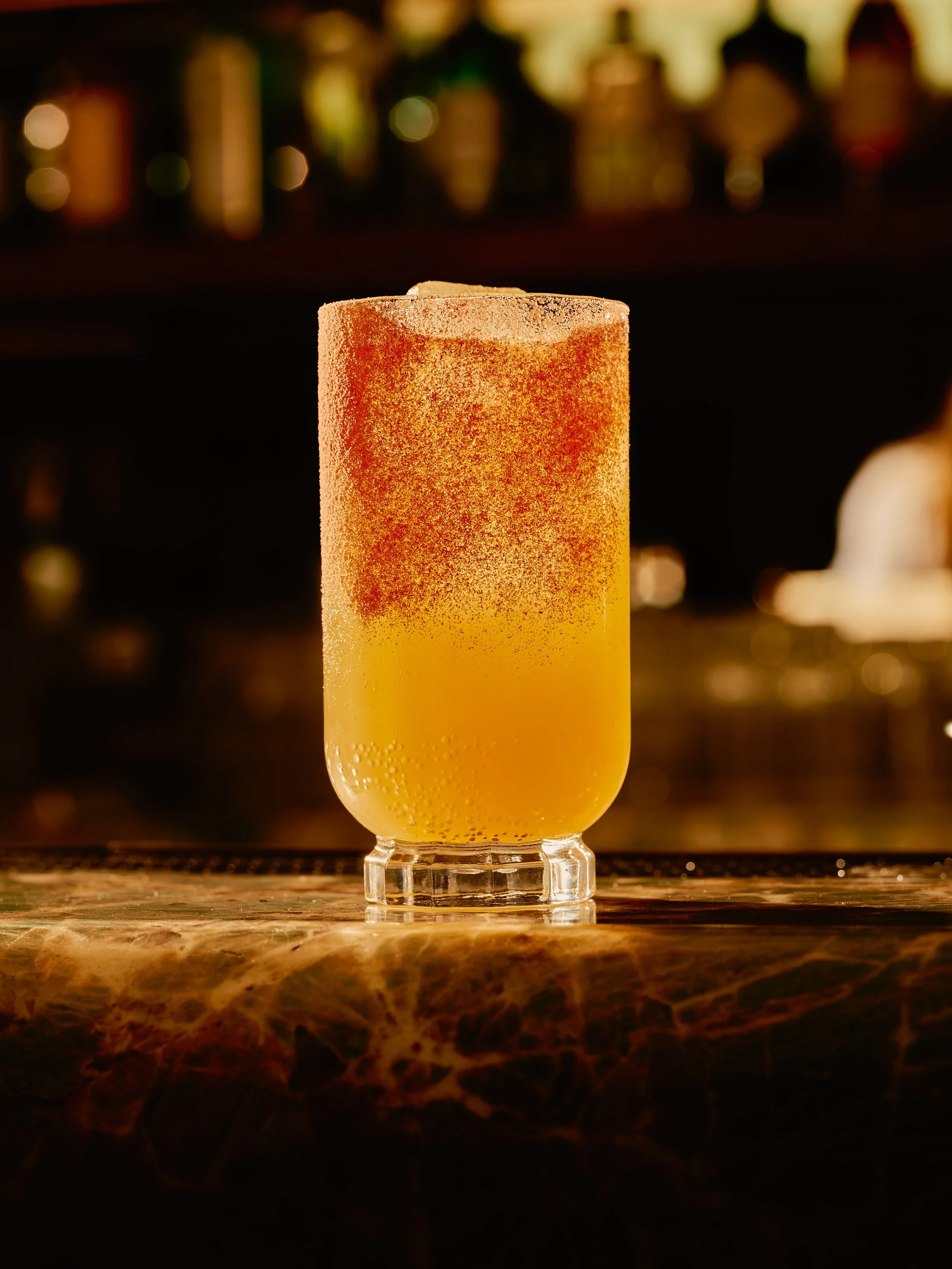 A tall, frosted glass filled with a yellow-orange beverage, topped with a salted rim, sitting on a marble bar counter with a blurred cityscape background.