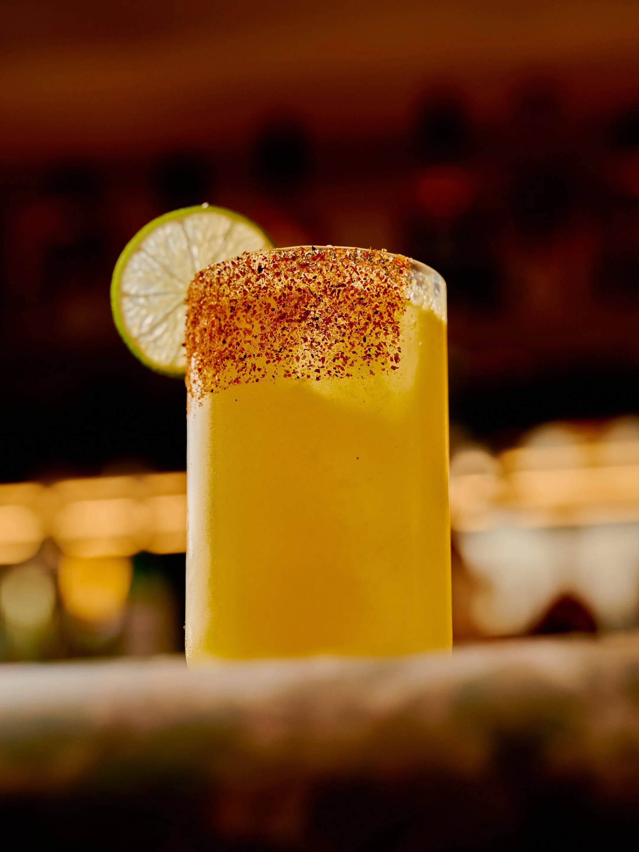 Close-up of a yellow cocktail garnished with a lime wedge and salted rim, with a blurred dark background.
