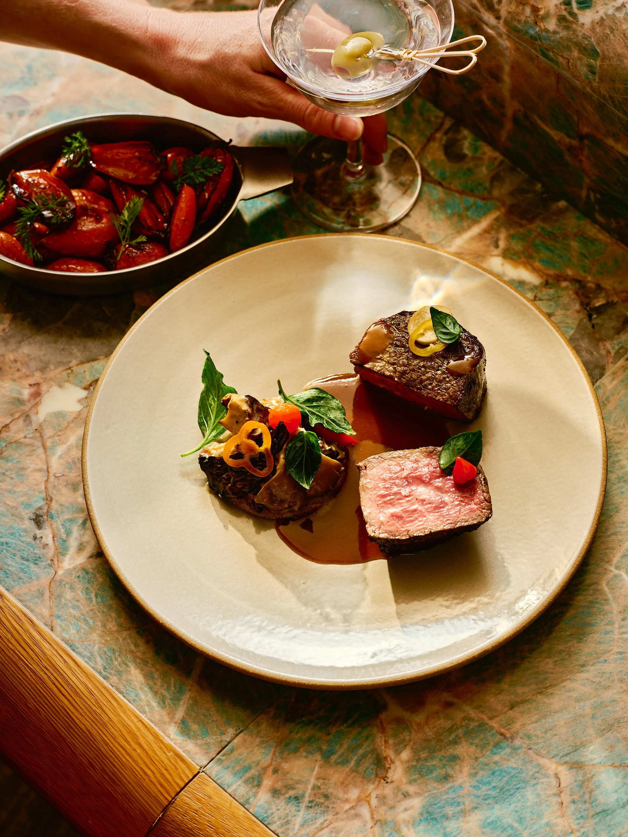 Plate with three pieces of cooked beef, garnished with herbs and vegetables, on a rustic table. In the background, a hand holds a glass of white wine and a bowl of strawberries.