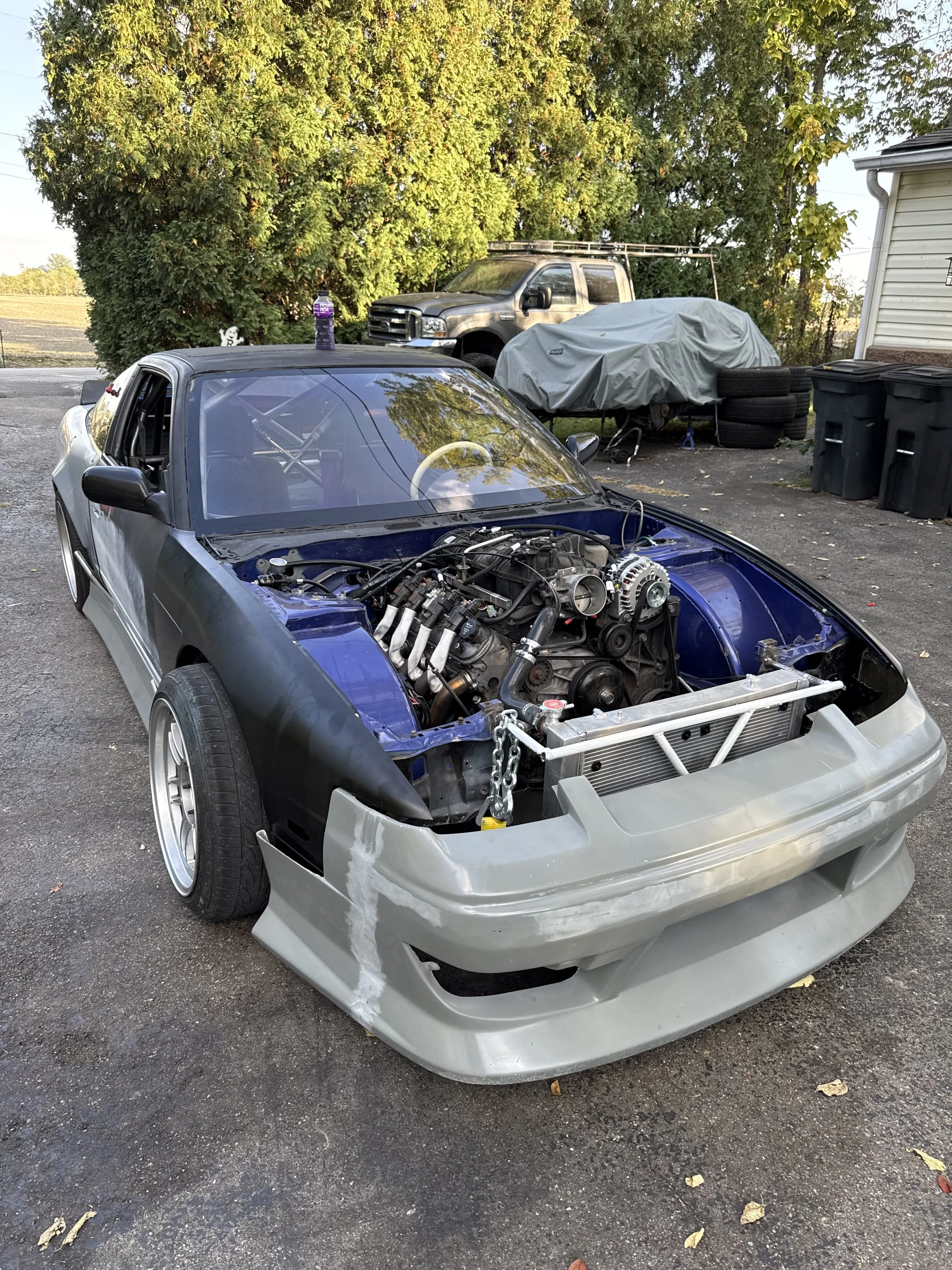 A partially assembled race car with an exposed engine bay, black front fenders, gray front bodywork, and silver rims parked on asphalt with trees and a house in the background.