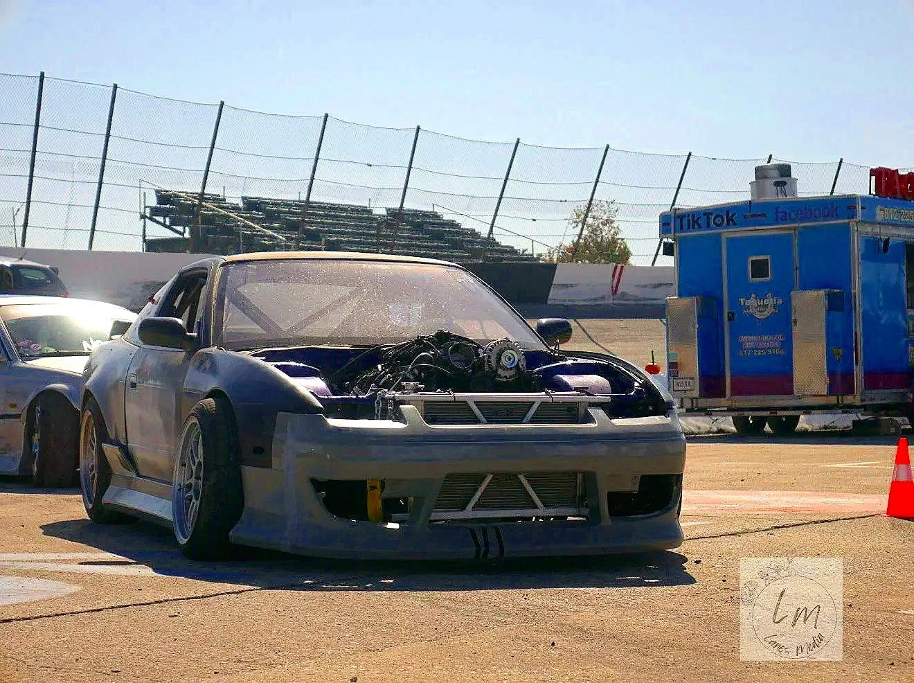 Race car on a pit lane with no front bumper, showing engine components, and another race car partially visible beside it, with a blue trailer in the background designed for social media platforms.