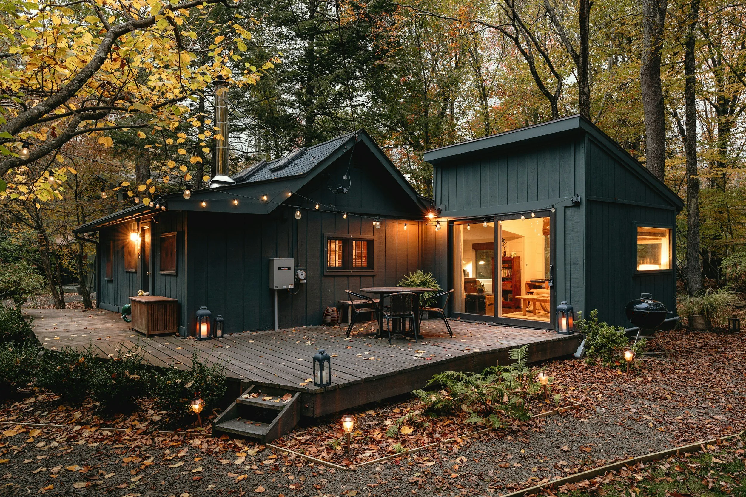 A cozy black house in the woods surrounded by fallen autumn leaves, with a wooden deck, outdoor string lights, lanterns, and a small grill, illuminated at dusk.