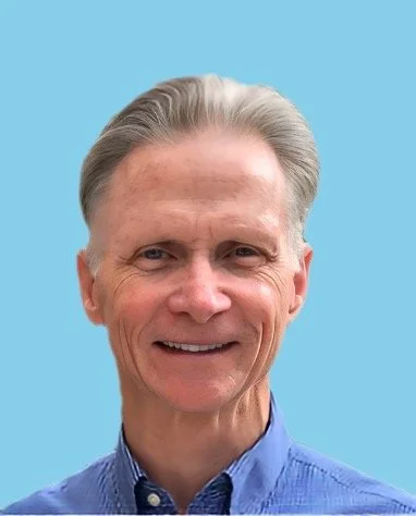 Portrait of an older man with gray hair wearing a blue collared shirt, smiling, against a light blue background.