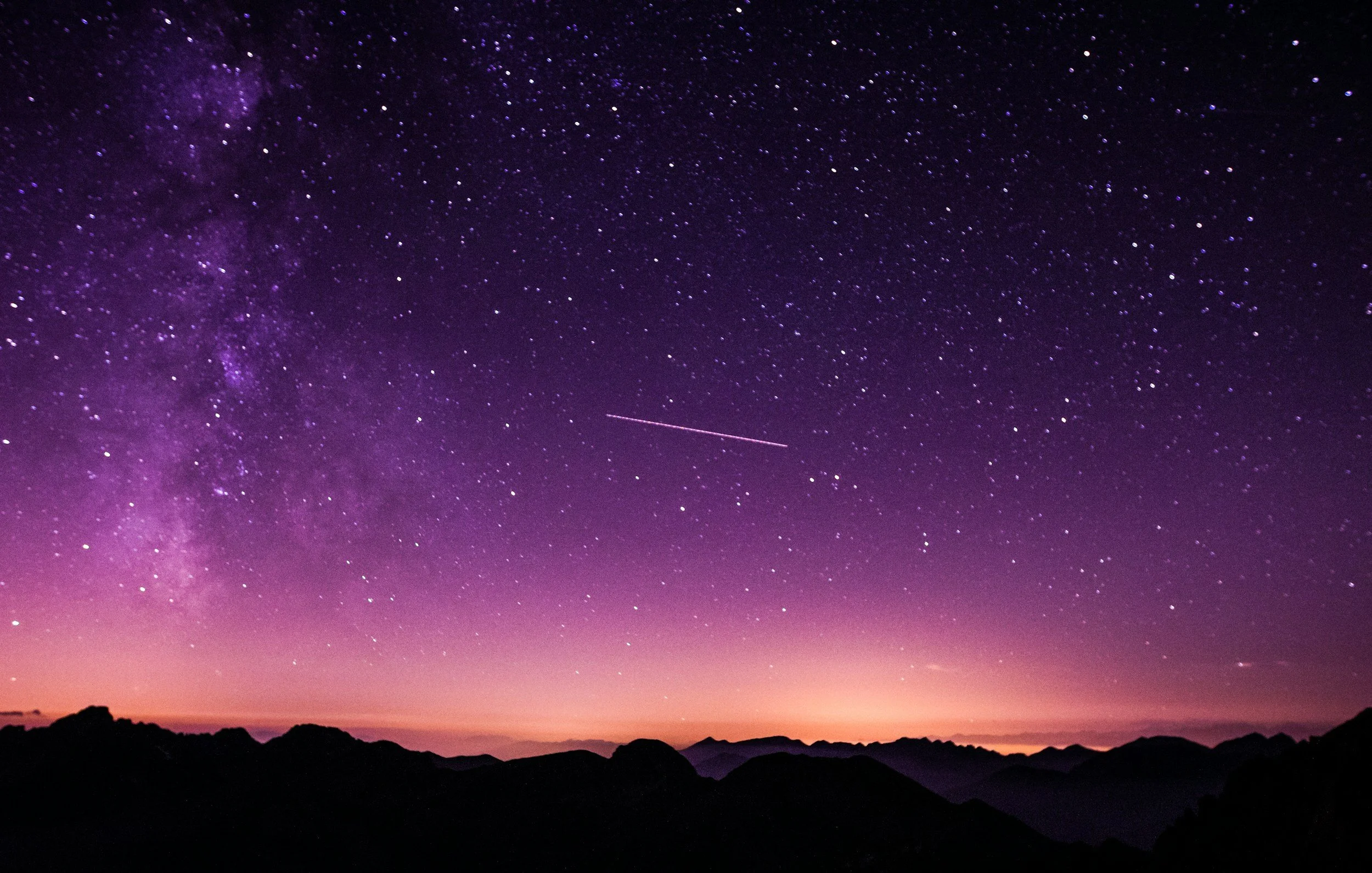 Night sky filled with stars and the Milky Way galaxy, with a streak of a shooting star, over silhouette of mountains.