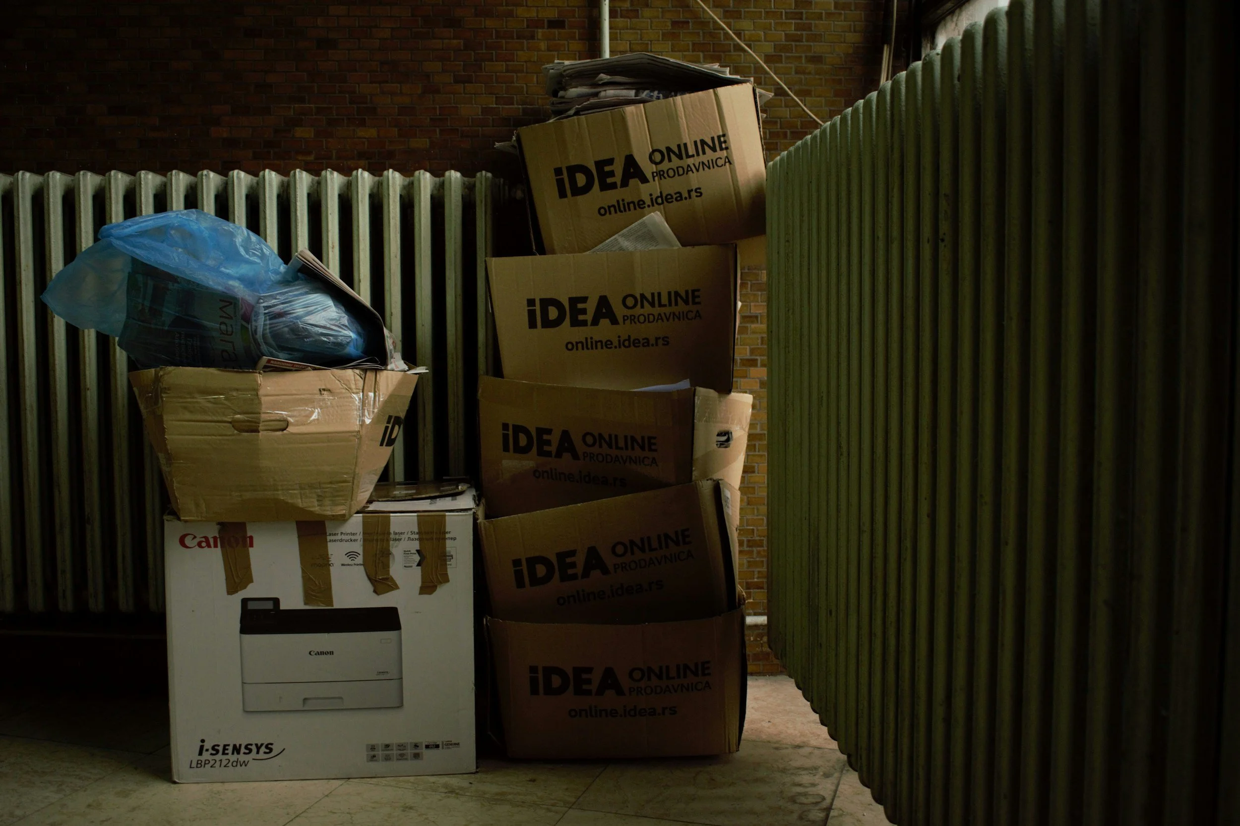 Stacked cardboard boxes and a Canon printer box beside a green corrugated fence and brick wall, with some boxes labeled 'IDEA ONLINE PRODAVNICA'.