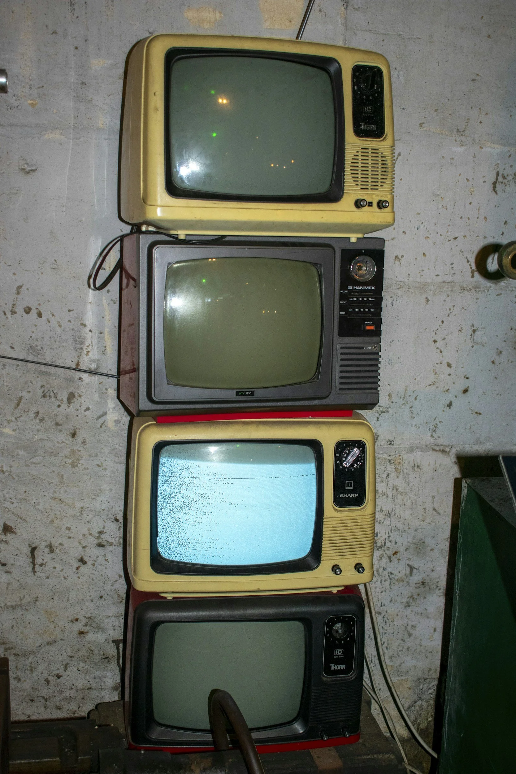Four vintage CRT televisions stacked vertically against a concrete wall. The top, third and bottom TVs are yellow, while the second has a gray frame. The bottom TV's screen is turned off, the middle TV displays static, the top TV has a few green and orange dots, and the third TV is on but its screen is blank.