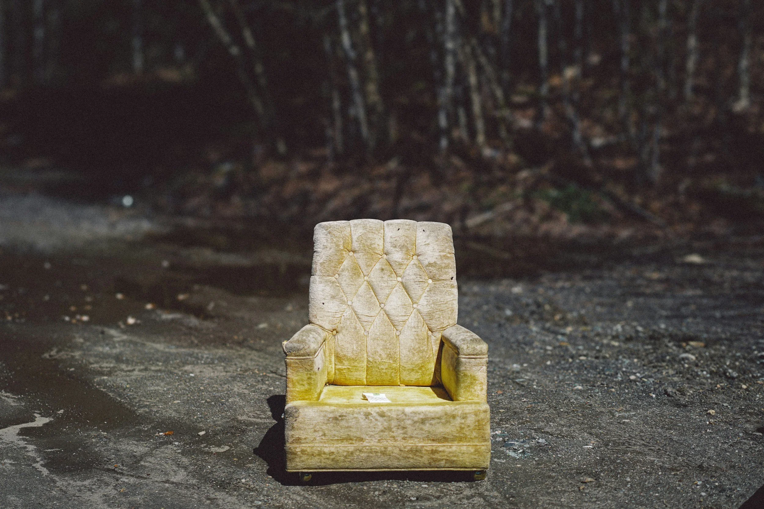 An old, yellowed, and worn armchair with a high backrest and padded seat placed outdoors on uneven dirt ground, with a dark forest background.