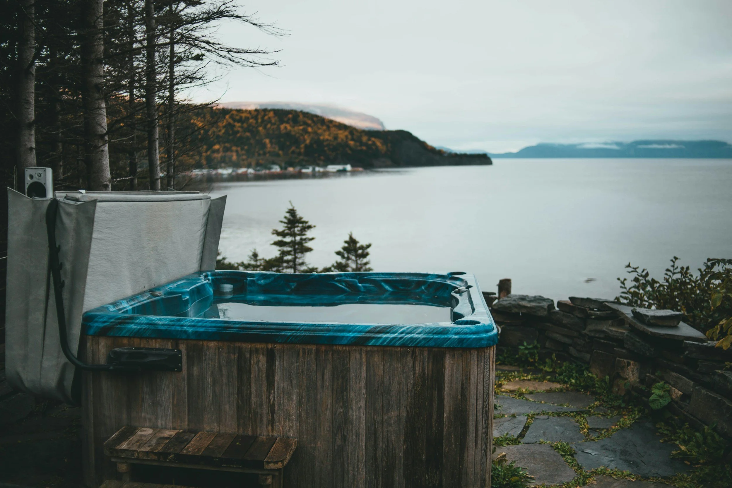 A hot tub filled with water on a patio overlooking a large lake, with trees and hills in the background during daytime.