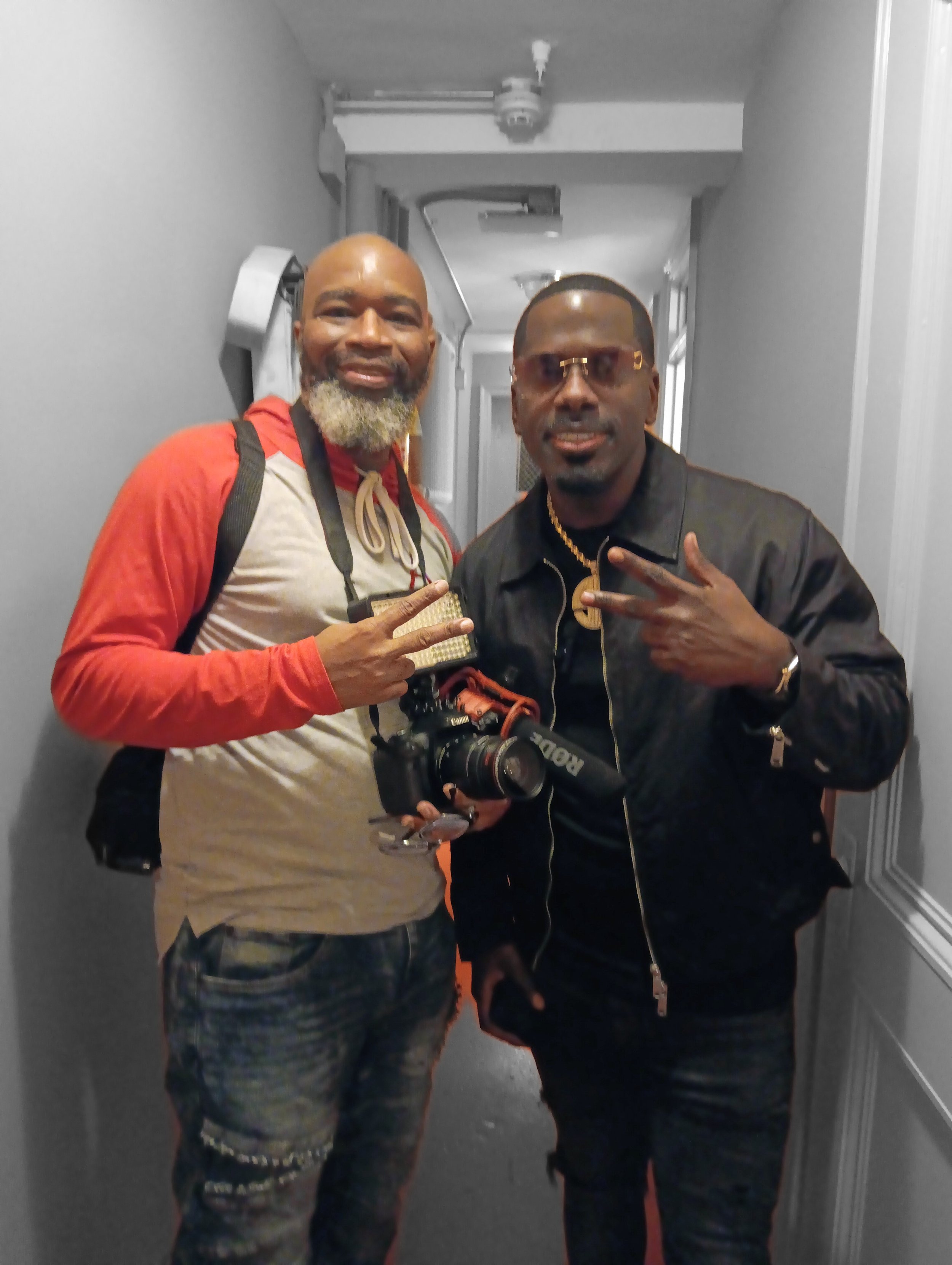 Two men standing in a hallway, one holding a camera, both flashing peace signs. The man on the left is wearing a red and beige hoodie with a backpack. The man on the right is wearing sunglasses and a black jacket with a camera strap around his neck.