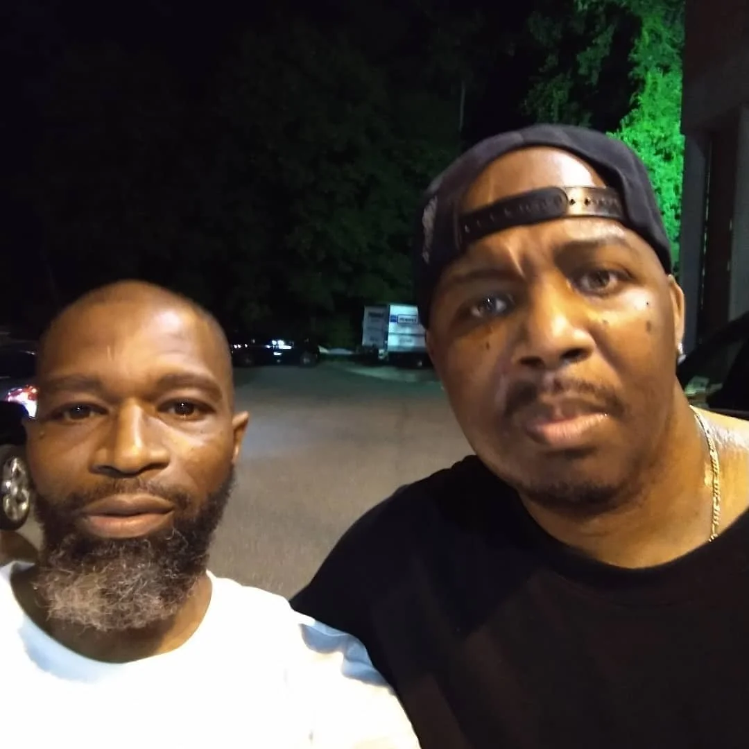 Two men taking a selfie outdoors at night, with trees and parked cars in the background.