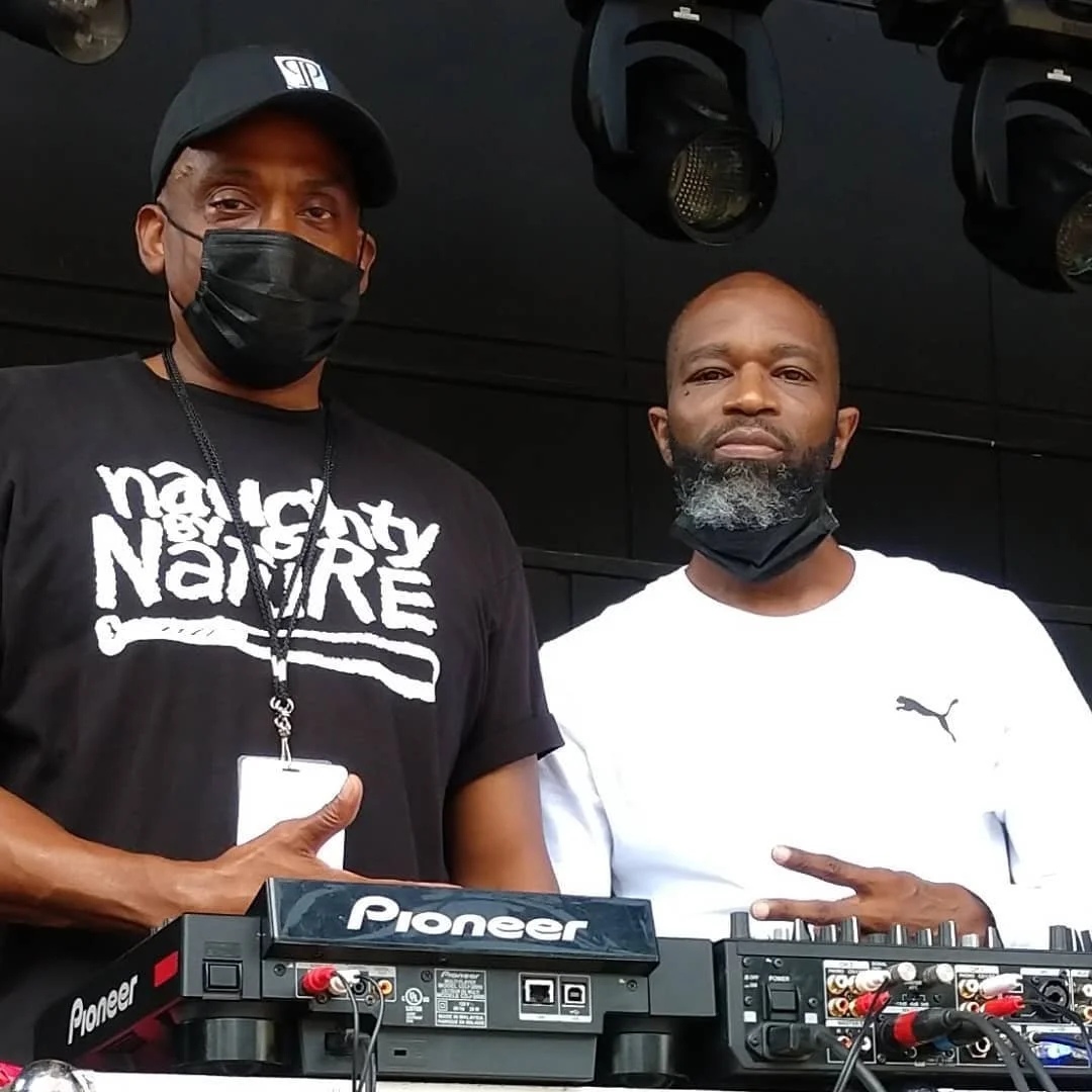 Two men standing behind DJ equipment, one wearing a black mask and a black cap, the other with a partially down face mask. Both are looking at the camera, with the man on the right making a peace sign. The equipment bears the brand name 'Pioneer'.