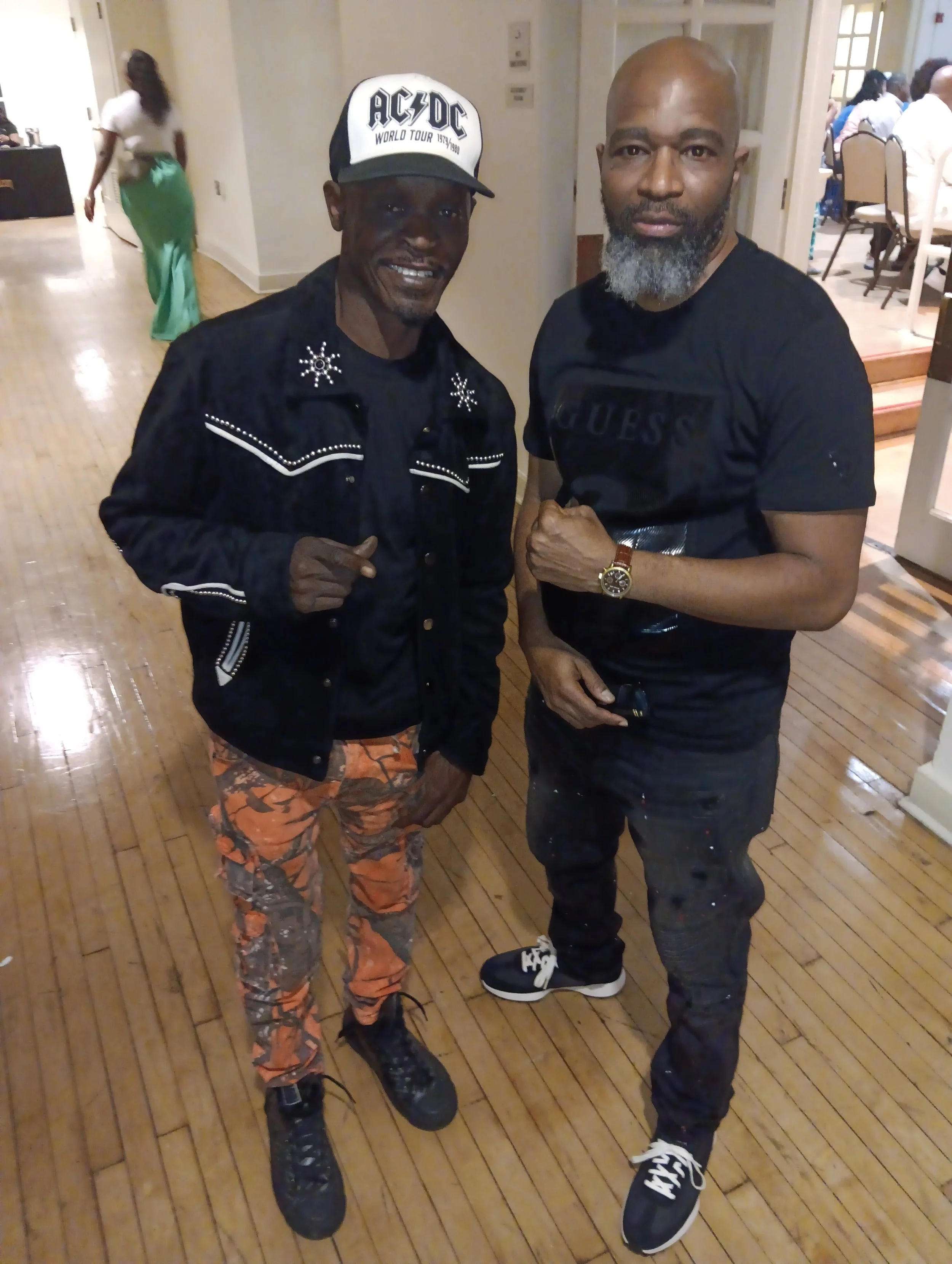 Two men standing indoors, one wearing a black jacket with white embellishments, orange and gray camouflage pants, and a black and white AC/DC cap, and the other in a black T-shirt with the GUESS logo, black pants, and sneakers. They are standing on a