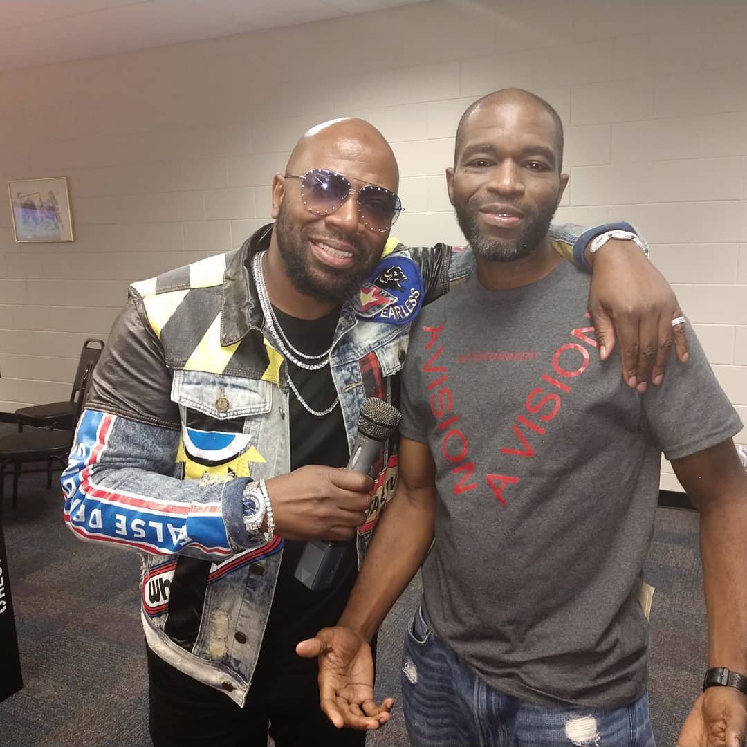 Me & Buckwild chillin, one with sunglasses and colorful jacket holding a microphone, and the other with a gray shirt with red text, in an indoor setting.