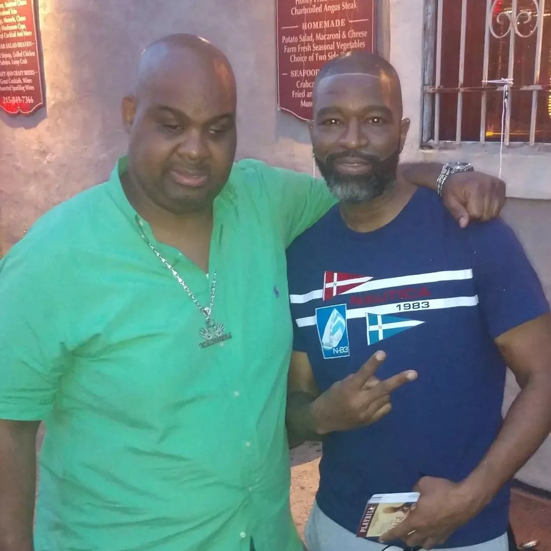 Two men standing side by side, smiling. One man is wearing a green shirt and the other is wearing a blue T-shirt with a graphic design. They are outside in front of a building with restaurant menu signs on the wall.