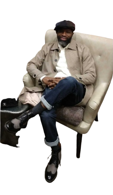 Tommy Slaughta sitting on a beige armchair with his legs crossed, wearing a gray jacket, white shirt, dark jeans, black shoes, and a black hat, smiling at the camera.