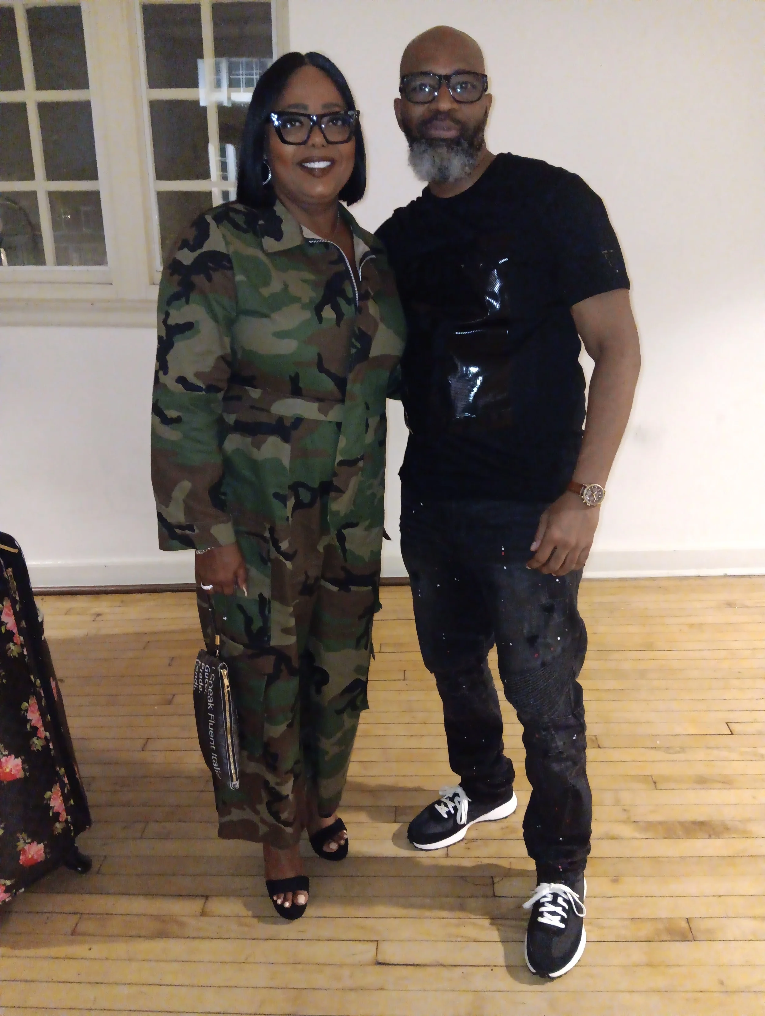 A woman and a man standing together indoors. The woman is wearing a camouflage suit, glasses, and black open-toe shoes. The man is dressed in black, wearing glasses, and black sneakers, with a beard and a watch.