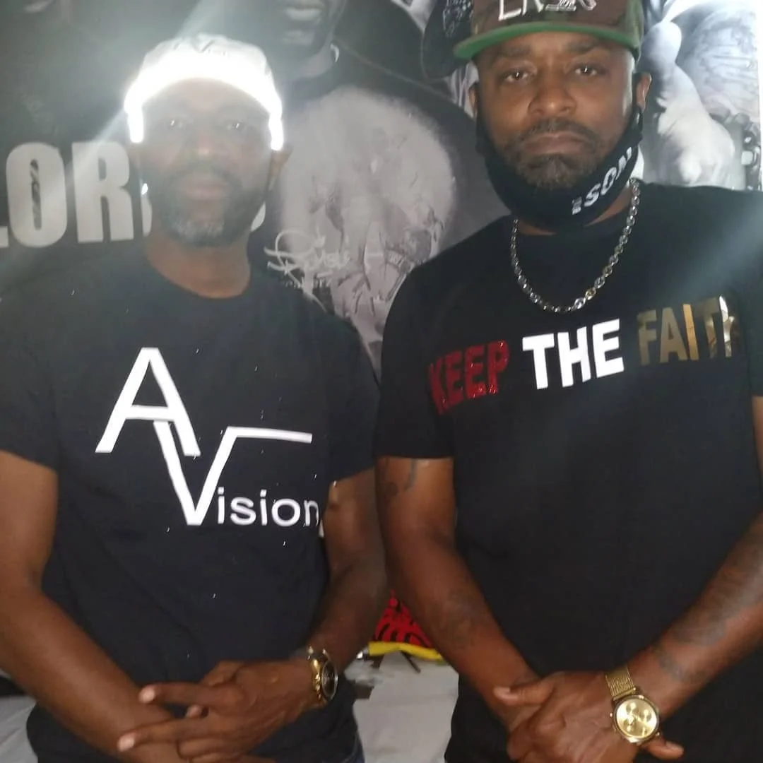 Two men standing side by side, both wearing black t-shirts with motivational messages, and accessories like watches and chains. They are in a dark environment with a graffiti-style background.