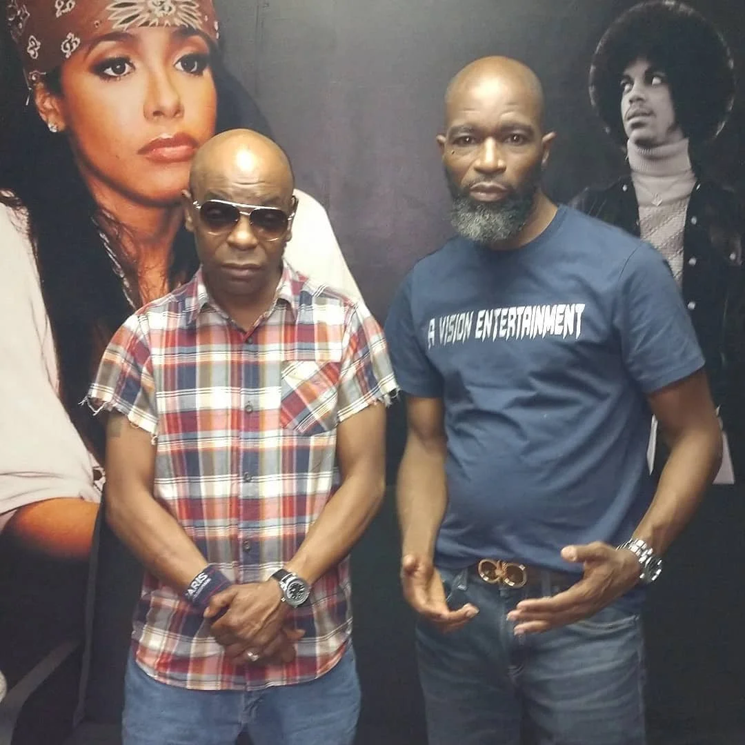 Two men standing in front of a backdrop with images of two women. The man on the left is wearing sunglasses and a plaid shirt. The man on the right is wearing a T-shirt that says 'A VISION ENTERTAINMENT'.