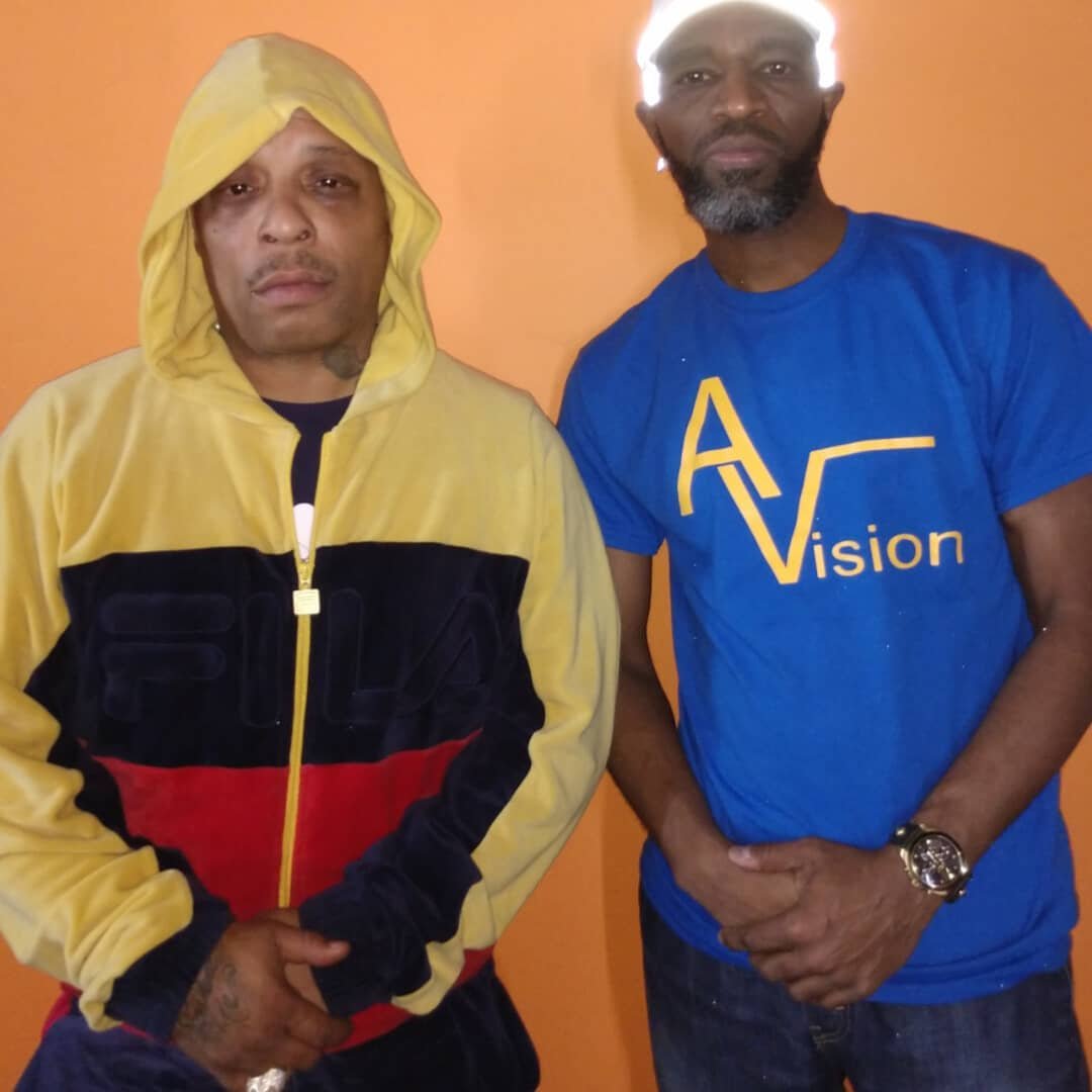 Two men standing against an orange wall, one wearing a yellow and black hoodie and the other wearing a blue T-shirt with 'AVision' written on it.