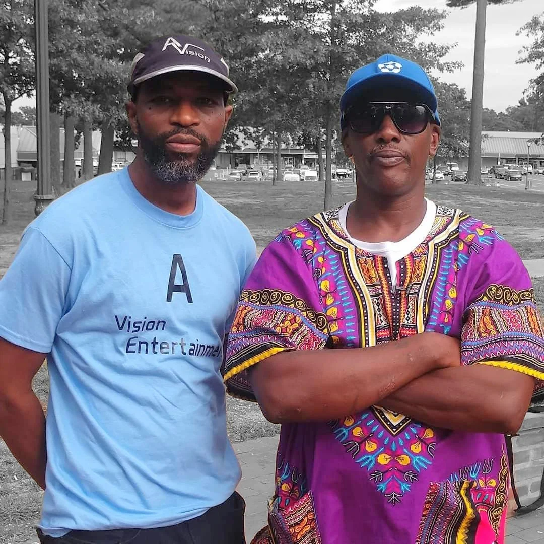 Two men standing outdoors, one wearing a black cap and light blue T-shirt with text, the other wearing a blue cap, sunglasses, and a colorful patterned shirt. Background features trees and buildings.