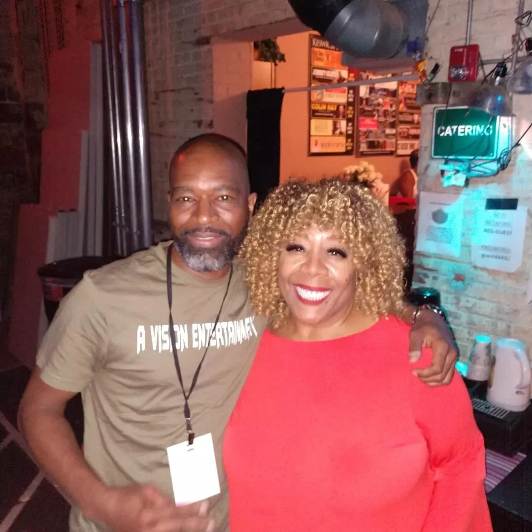 A man and woman smiling and posing together in a restaurant or bar, with the man wearing a lanyard and the woman wearing a red top, in a cozy industrial-style setting with brick walls and a "Catering" sign.